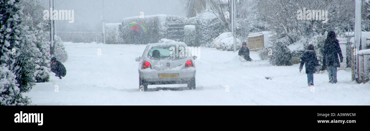 Route de village avec la neige qui tombe les enfants jouant personnes à pied et en voiture avec feux arrière sur Banque D'Images
