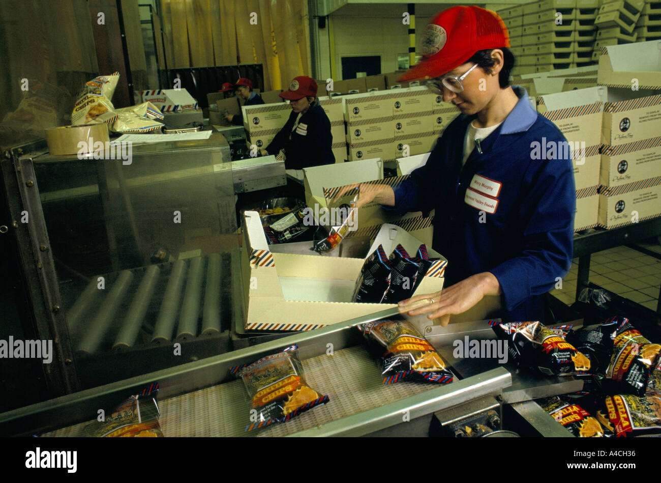 OUVRIER D'USINE EMBALLANT DES PAQUETS DE PUCES DE TORTILLA DE LA CEINTURE DE CONVEYOT DANS LA BOÎTE, UKG Banque D'Images