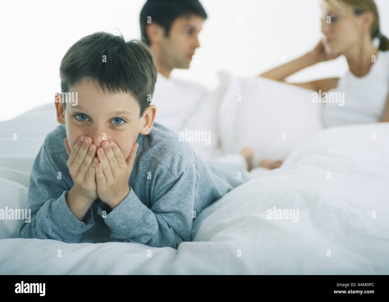 Boy lying on stomach in bed couvrant la bouche avec les mains, les parents de discuter en arrière-plan Banque D'Images