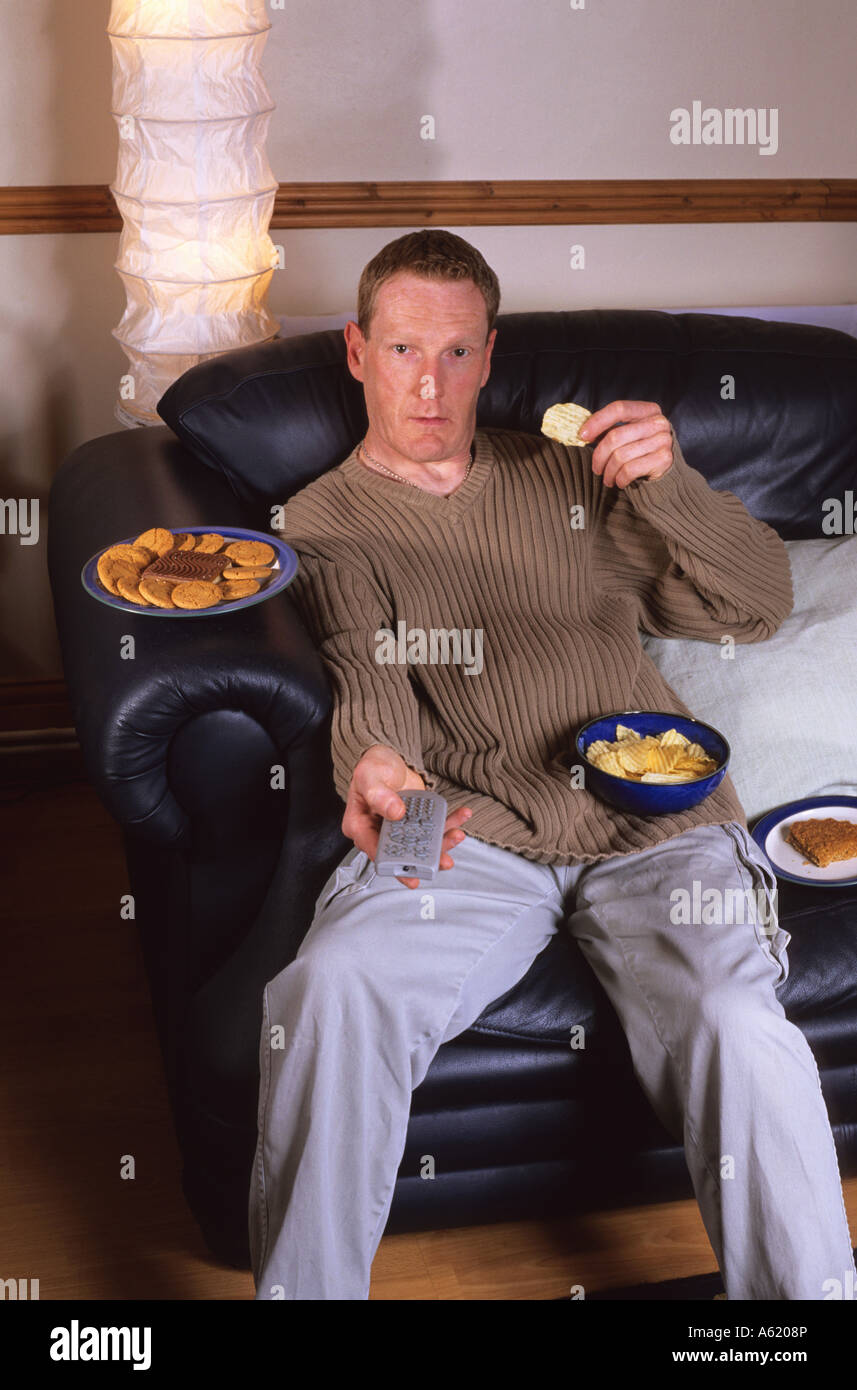 Homme assis sur setee entouré de junk food holding television remote controller Banque D'Images