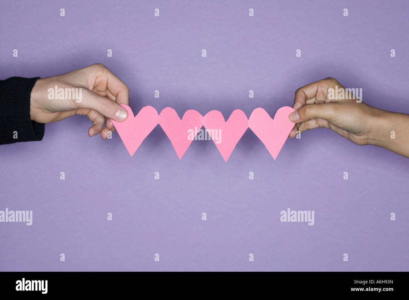 Close up of mixed race deux mains tenant du papier rose découper des cœurs en studio entre eux en fond violet. Banque D'Images