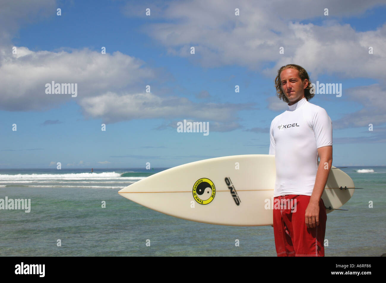 Homme en blanc rash guard holding surfboard contre partiellement nuageux ciel et les vagues de l'océan en arrière-plan Honolulu Oahu Hawaii Banque D'Images