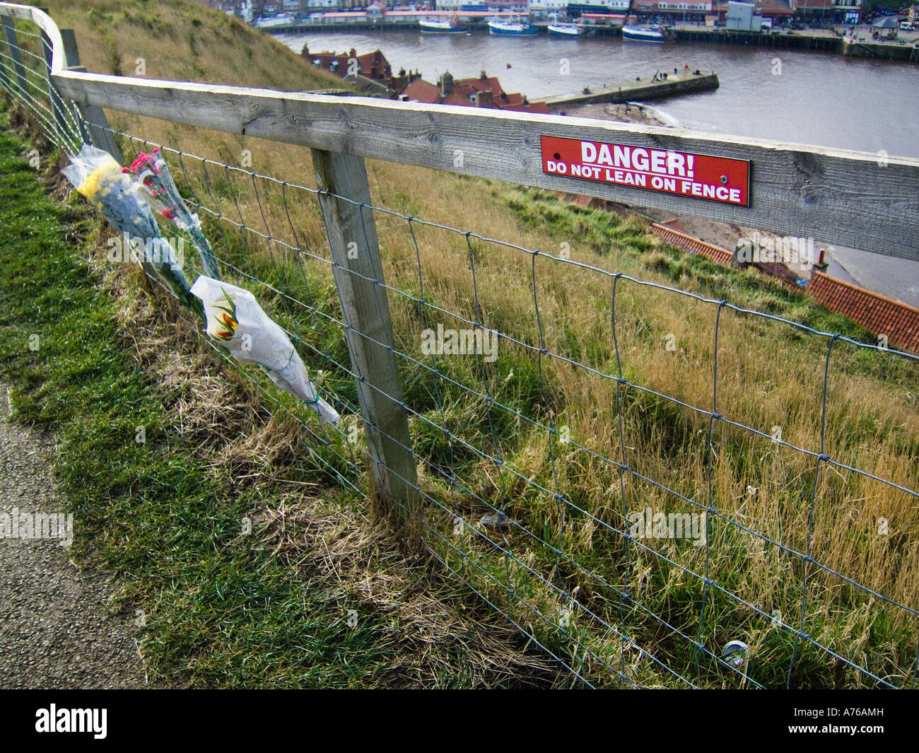 Dangerous Cliffs Do Not Lean on Fence signe sur une clôture en treillis métallique en haut d'une falaise, à côté de bouquets de fleurs attachés à une clôture dans un acte de souvenir Banque D'Images