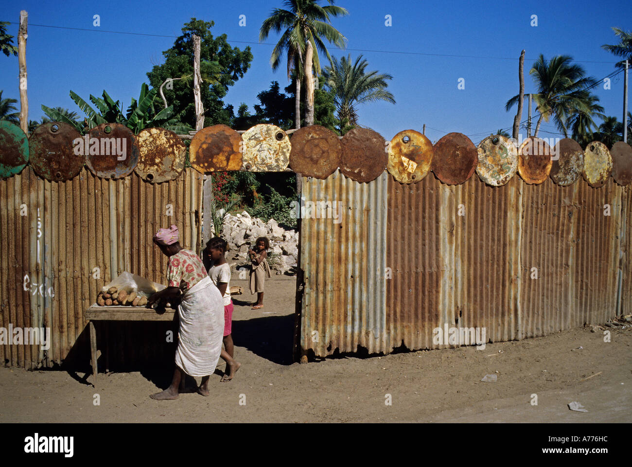 Femme en costume traditionnel vend du pain dans la rue avec clôture en tôle ondulée et le tambour à huile couvercles dans le sud de remorquage malgache Banque D'Images