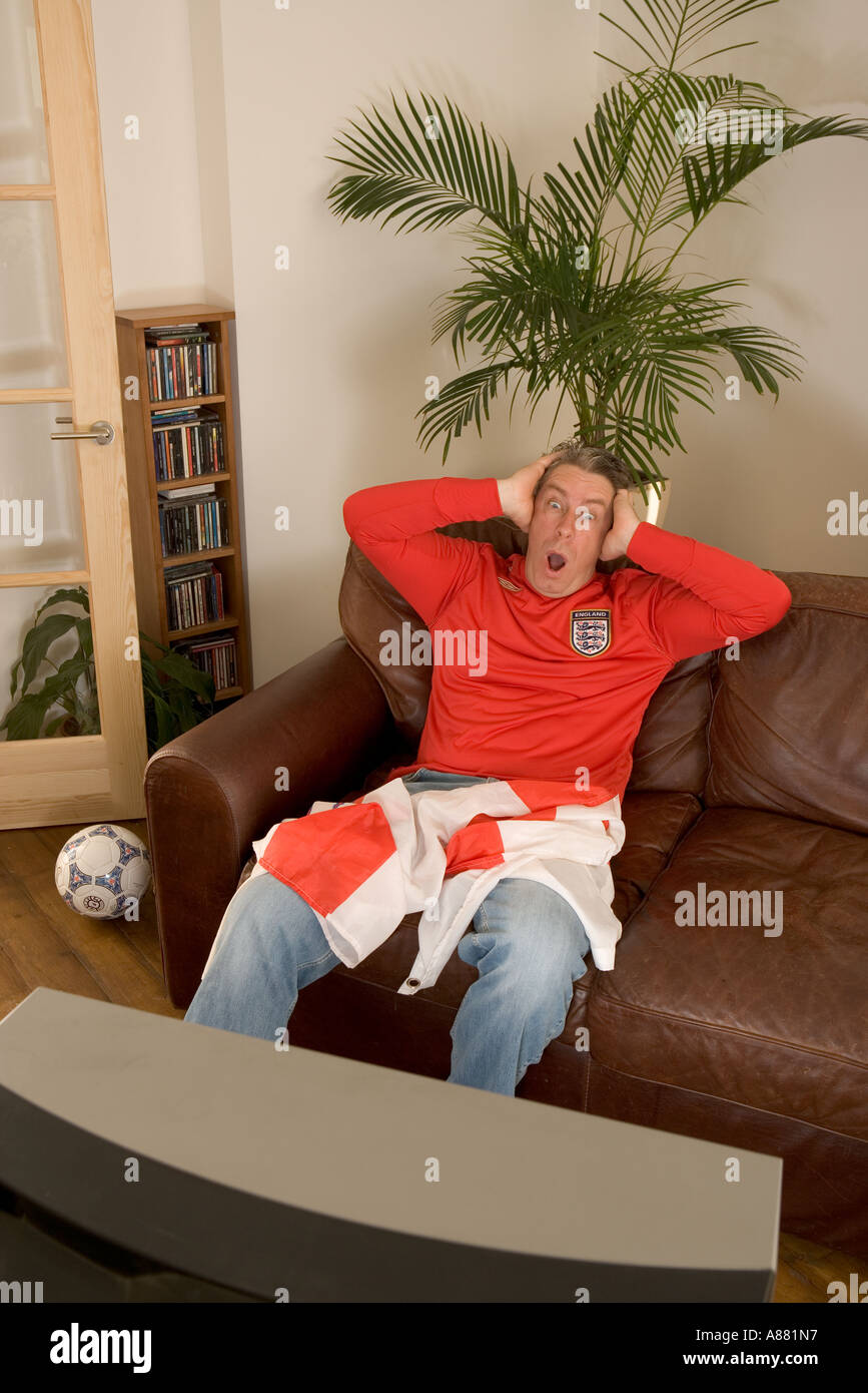 Parution du modèle. Angleterre football fan assis à la maison sur le canapé devant correspondre à la télévision déçu et contrarié que perd l'équipe. Banque D'Images