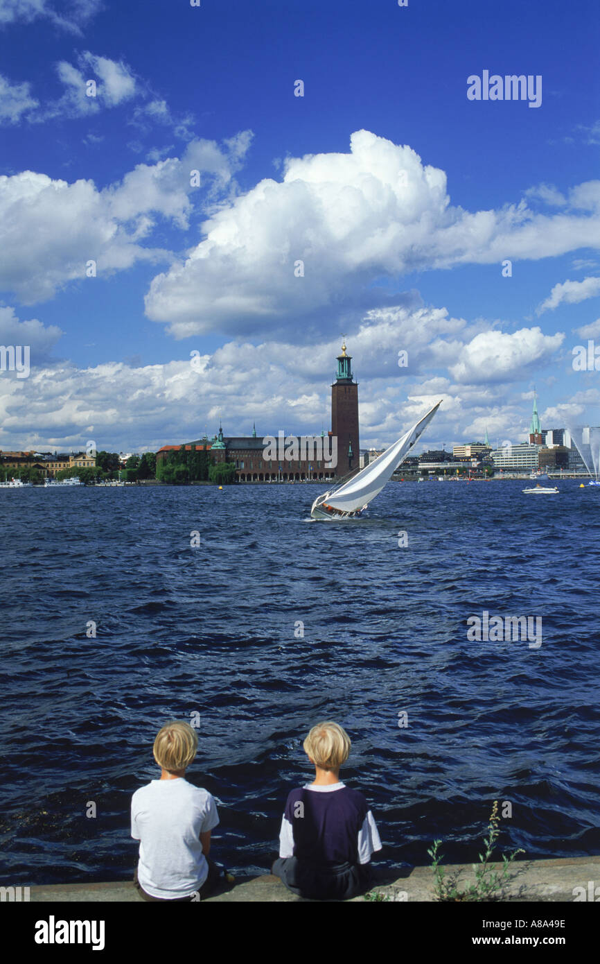 Deux garçons assis sur les eaux de Riddarfjarden avec voilier et l'Hôtel de Ville de Stockholm Banque D'Images