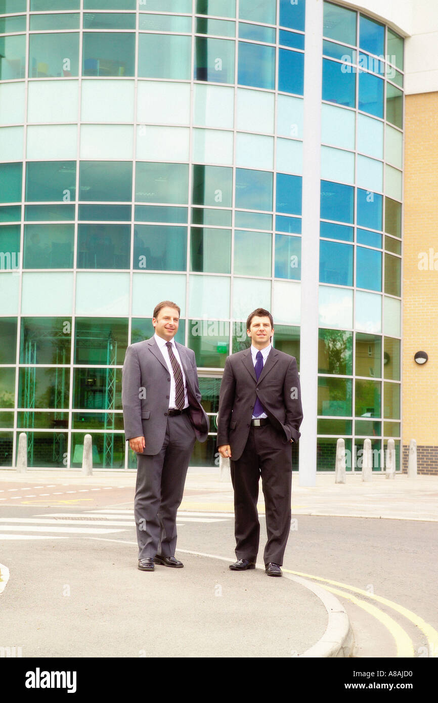 Deux hommes d'affaires caucasiens en costumes debout à l'extérieur d'un immeuble de bureaux moderne avec une façade vitrée courbée Banque D'Images