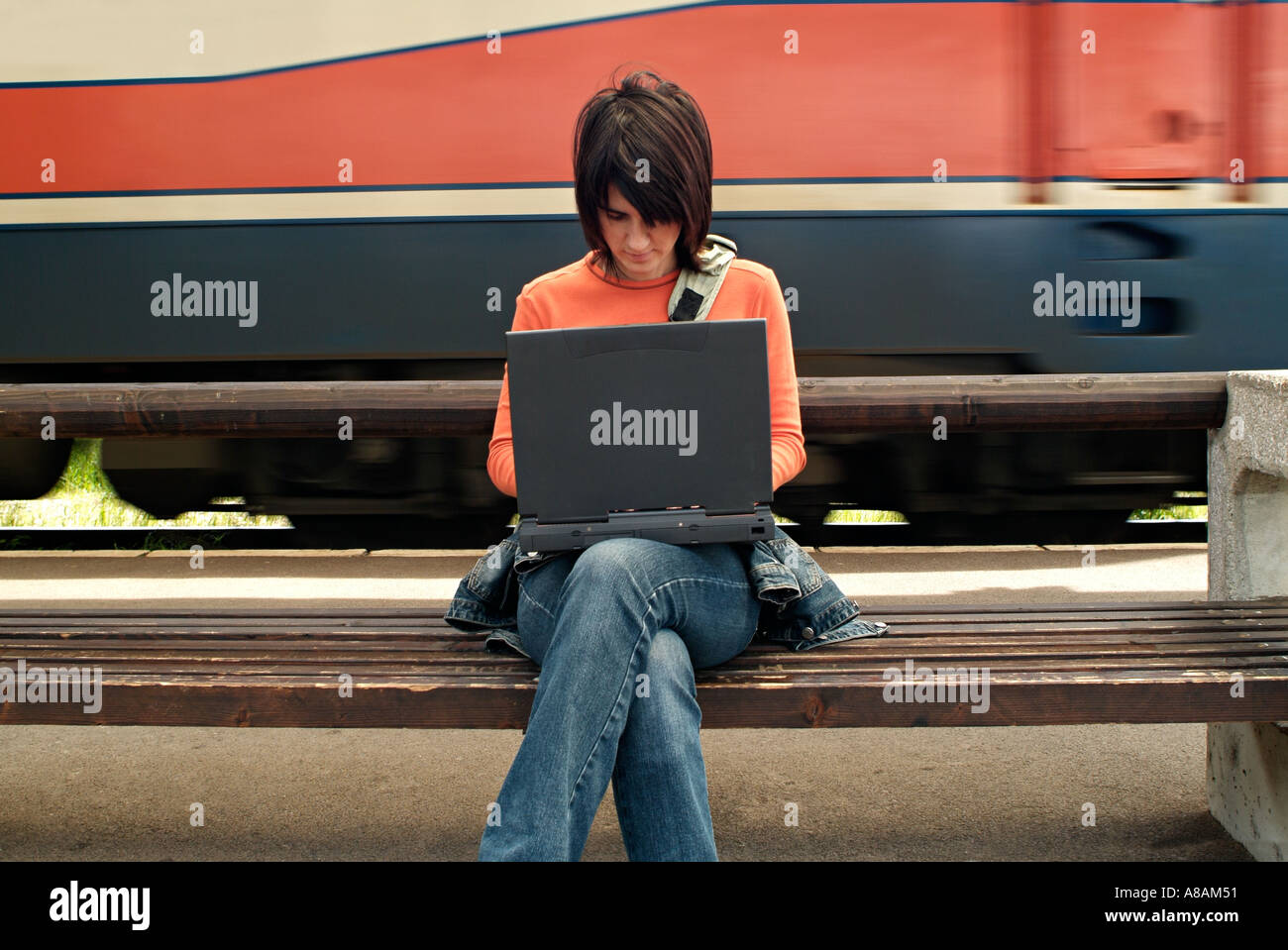 Jeune femme assise sur un banc sur une plate-forme de la gare de la saisie sur un ordinateur portable comme un train passe Banque D'Images
