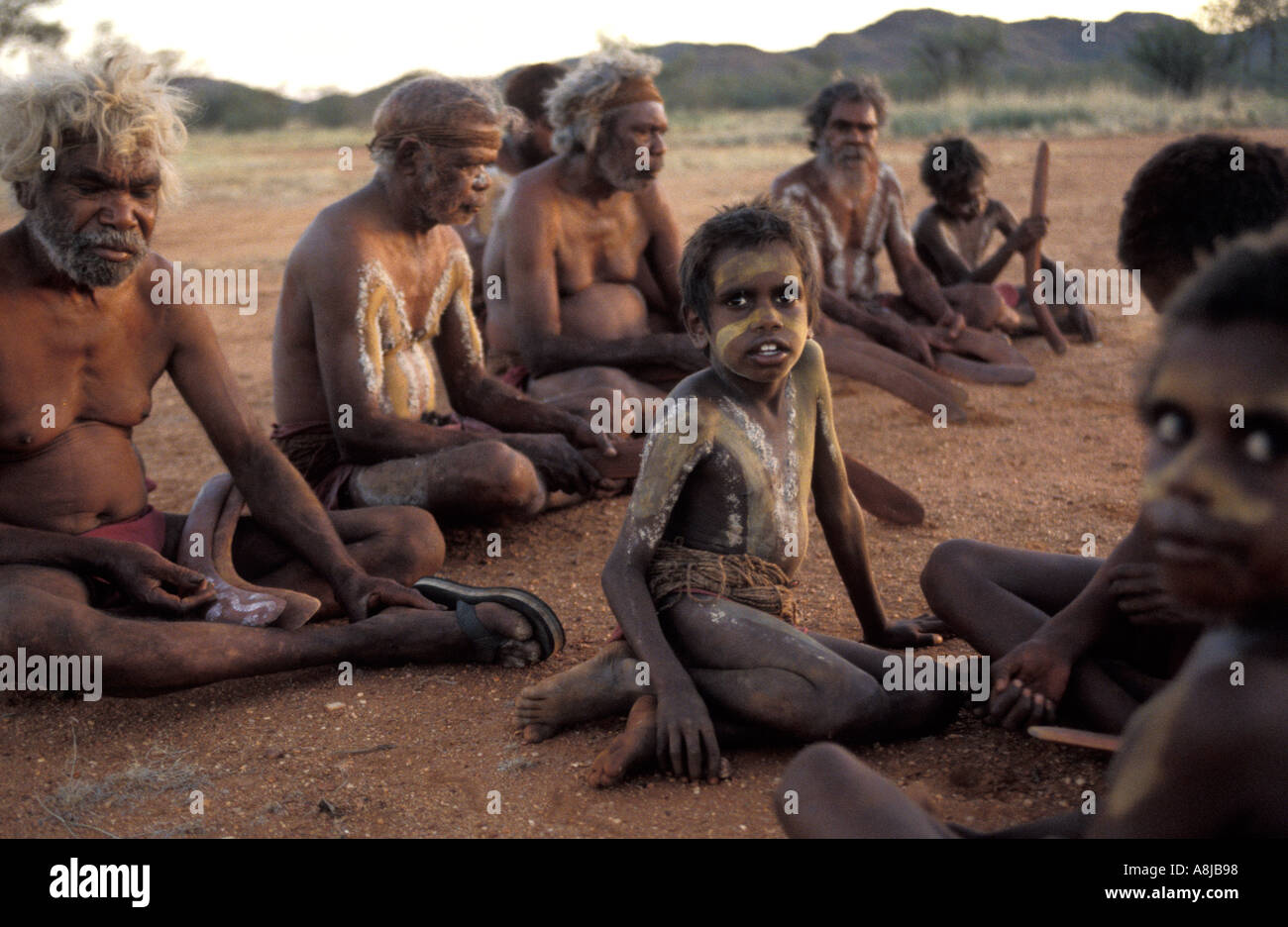 Les aînés autochtones et les jeunes garçons dans la peinture du corps de cérémonie s'asseoir sur le sable rouge par le feu après le coucher du soleil dans le désert de l'Australie centrale Banque D'Images