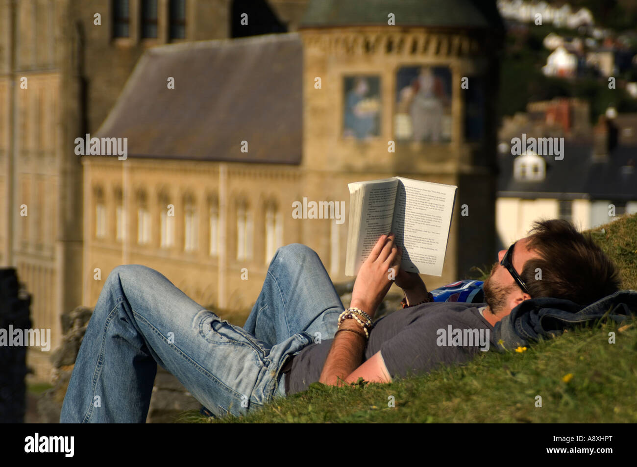 L'homme allongé sur le dos sur l'herbe la lecture d'un livre avec l'University of Wales Aberystwyth Ancien Collège en arrière-plan, l'été Banque D'Images