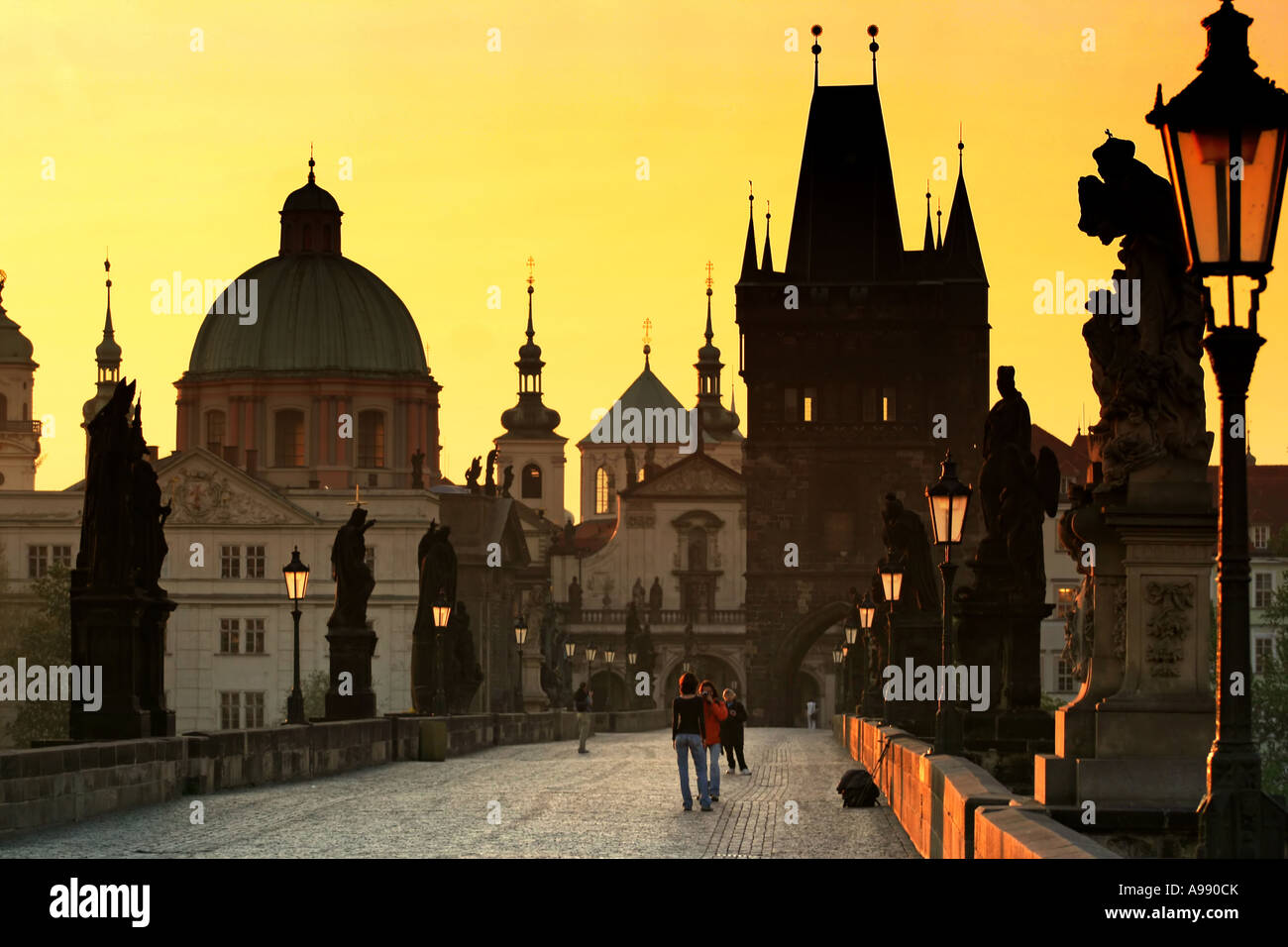 L'heure d'or baigne l'emblématique pont Charles de Prague dans une lueur chaleureuse, tandis que des statues aux silhouettes montent la garde sur la scène matinale sereine Banque D'Images