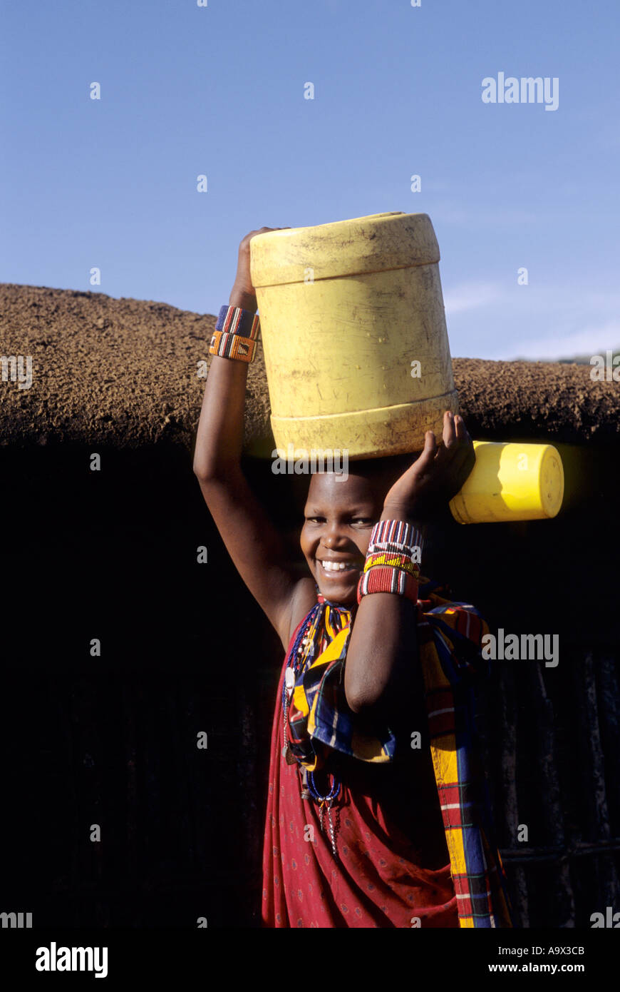 Lolgorian, au Kenya. Manyatta Masaï Siria ; sourire à l'extérieur d'un Massaï maison fortifiée de boue portant un baril de plastique jaune sur la tête. Banque D'Images