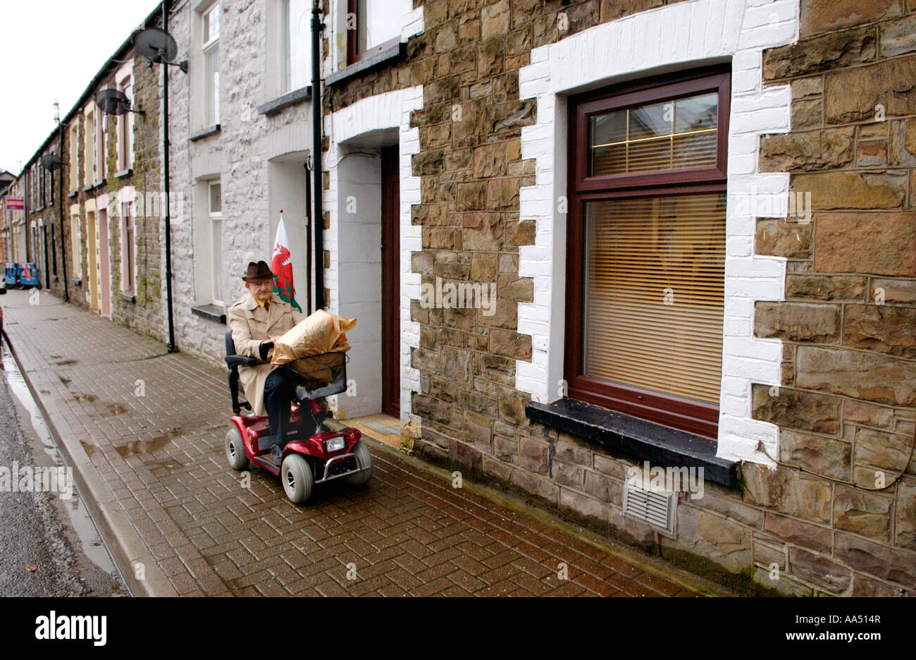 Vieil homme sur la mobilité scooter flying dragon rouge d'un drapeau tandis que dehors sur la rue commerçante de Treorchy, Valley South Wales UK Banque D'Images