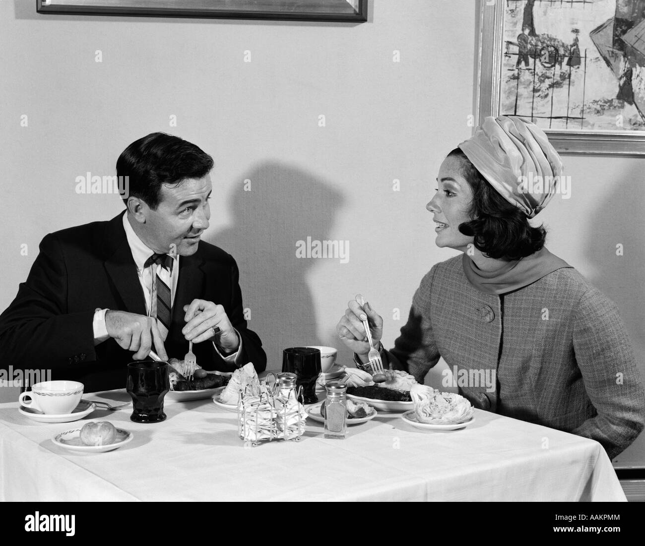 1960 HOMME FEMME COUPLE EATING MEAL IN RESTAURANT FEMME PORTE STYLE TURBAN HAT Banque D'Images