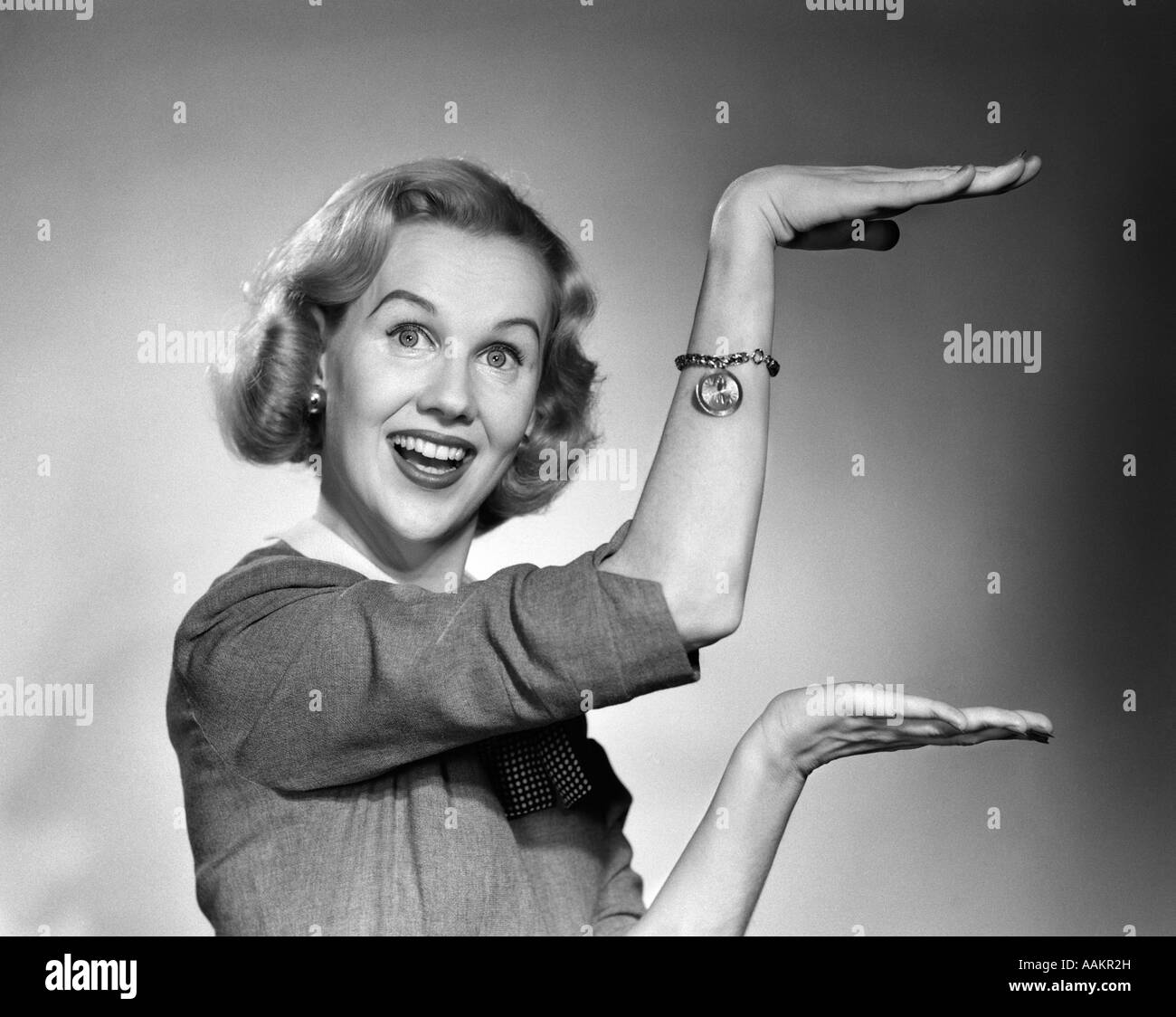 Années 1950 Années 1960 HAPPY SMILING BLONDE WOMAN GESTURING WITH HANDS MONTRANT LA TAILLE DE QUELQUE CHOSE LOOKING AT CAMERA Banque D'Images