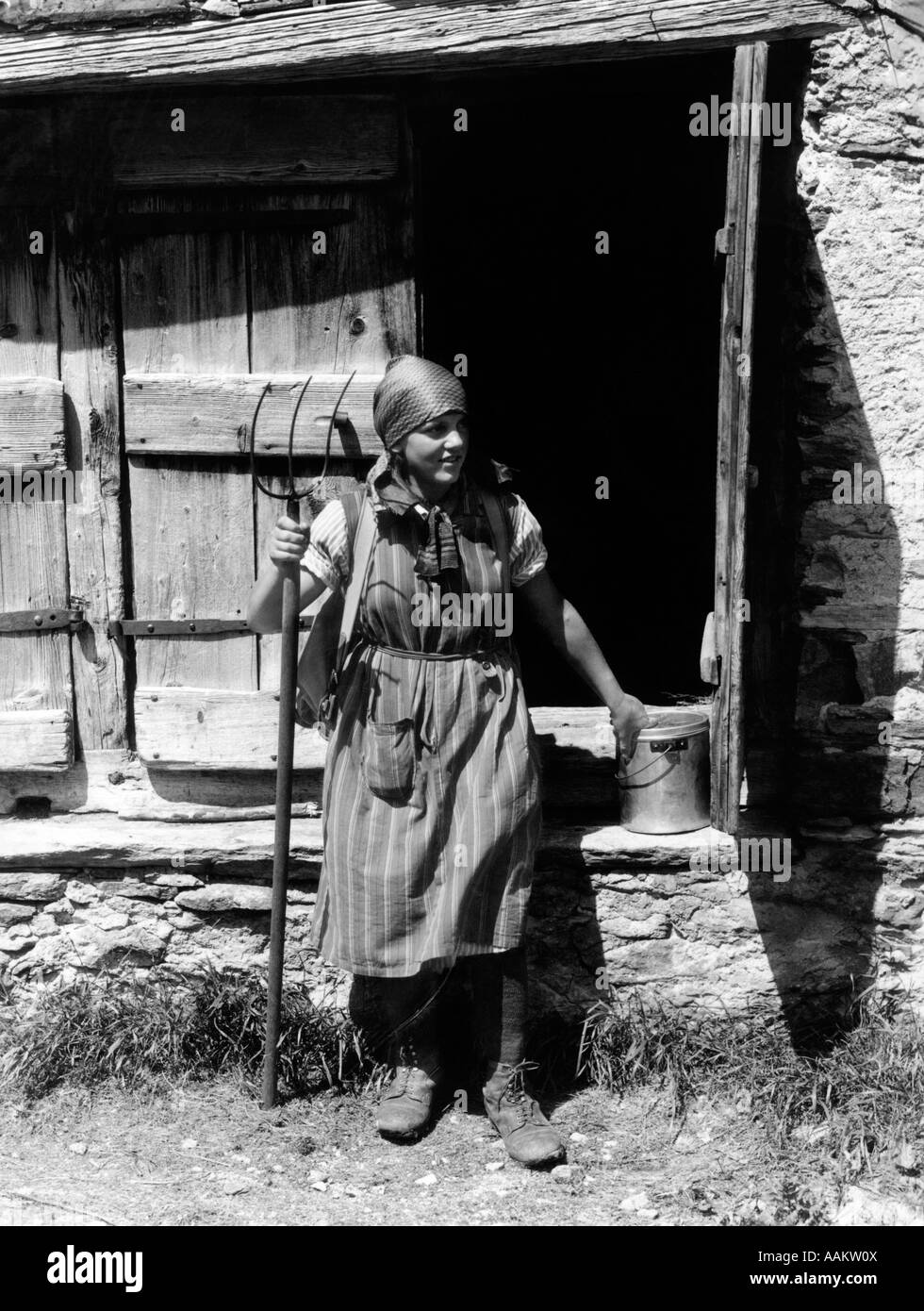 1930 FEMME EN COSTUME DE L'ESPACE RURAL SUISSE HOLDING PITCHFORK ET PAN EN FACE DE BÂTIMENT DE FERME EN PIERRE PRÈS DE LA FENÊTRE DE LA FURKA Banque D'Images