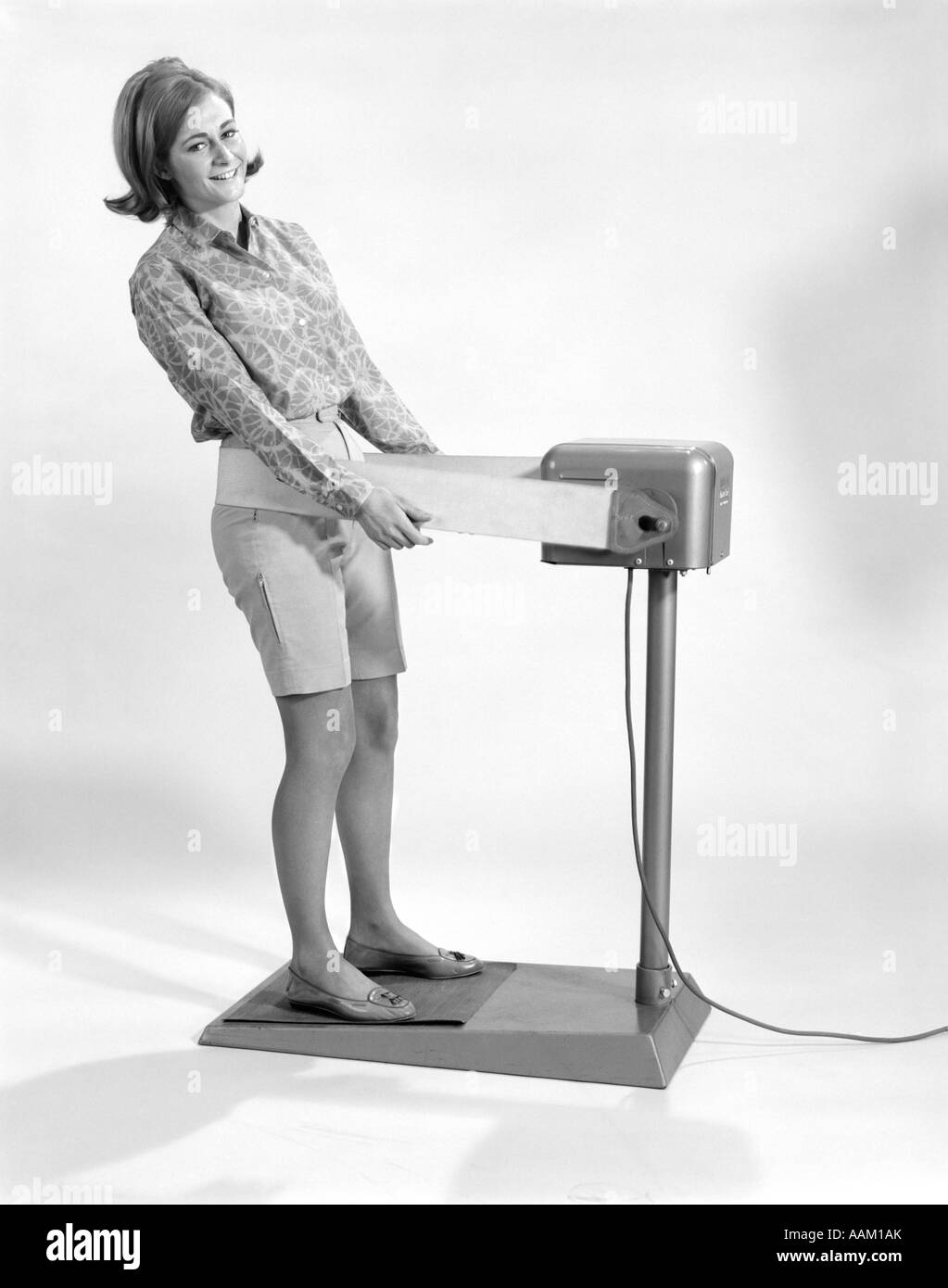 1960 SMILING YOUNG WOMAN STANDING SUR LA RÉDUCTION DE POIDS MACHINE D'EXERCICE VIBRANT LOOKING AT CAMERA Banque D'Images