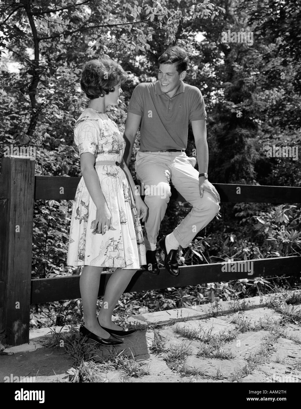 1960 TEENAGE COUPLE TALKING FLIRT DATING YOUNG WOMAN STANDING YOUNG MAN SITTING ON FENCE Banque D'Images