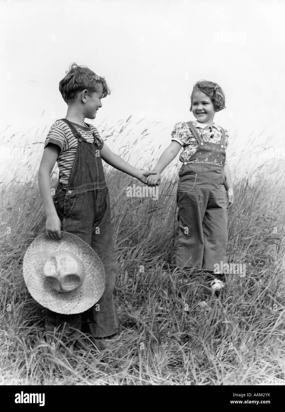Années 1930 Années 1940 FARM KIDS BOY GIRL WEARING BRETELLES tenant la main  descendre la colline herbeuse BOY CARRYING STRAW HAT Photo Stock - Alamy