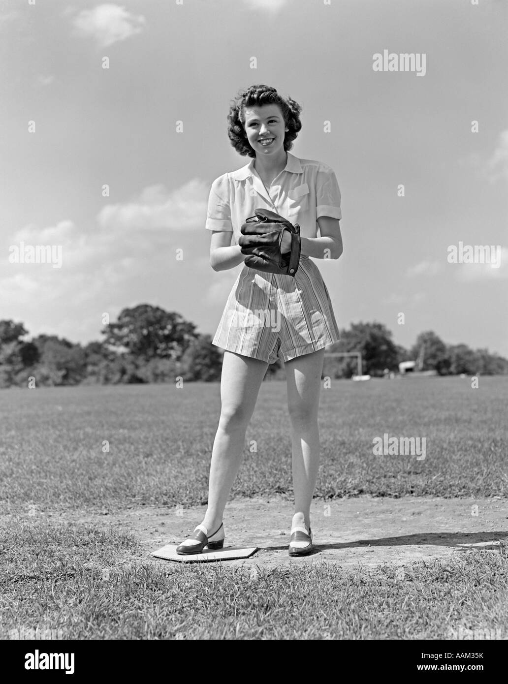 1940 SMILING TEEN GIRL PORTANT DES SHORTS DEBOUT SUR PITCHERS MOUND SOFT BALL GLOVE MITT SUR SA MAIN LE SOFTBALL Banque D'Images