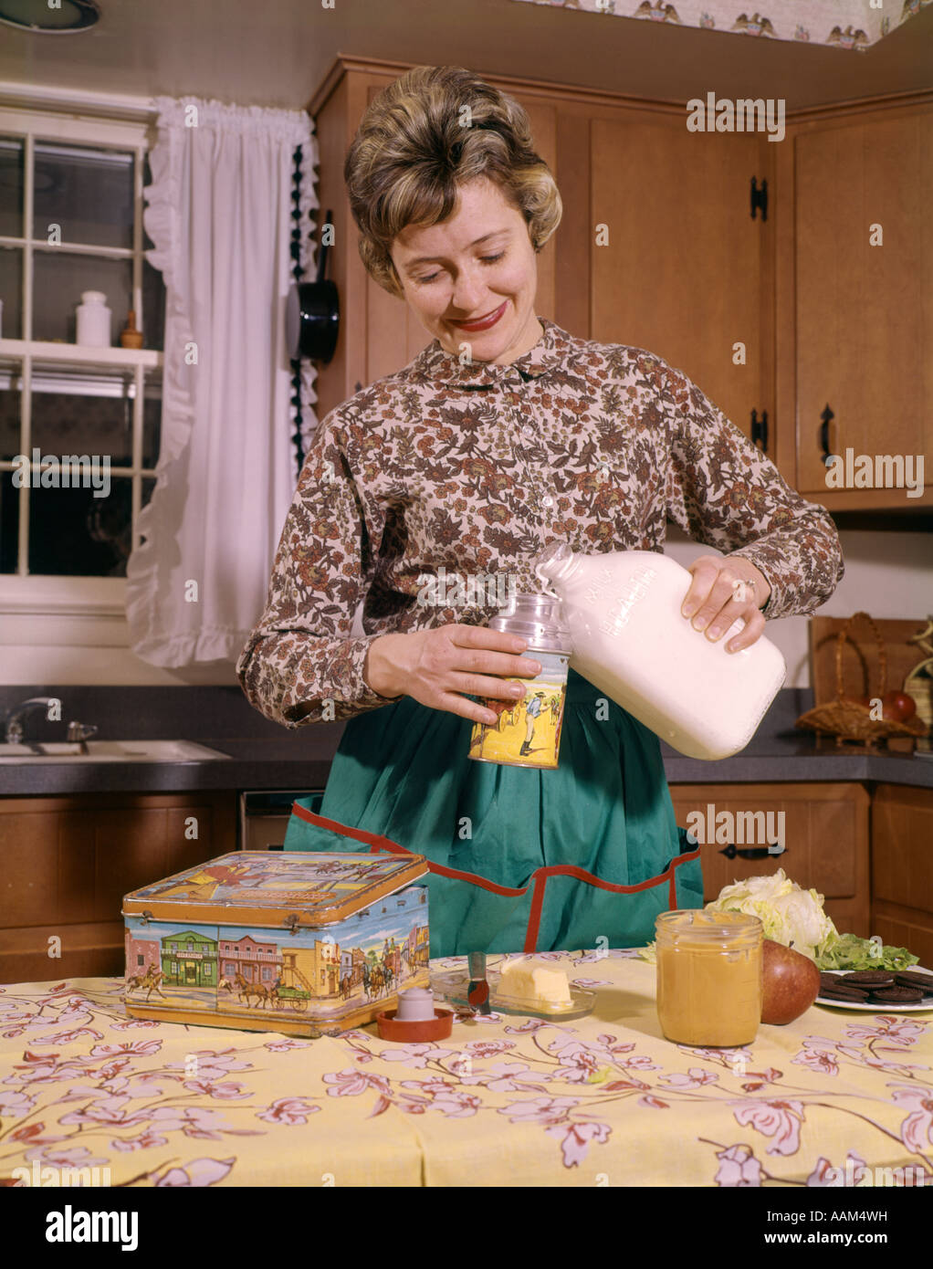 1960 SMILING WOMAN femme au foyer mère portant un tablier de cuisine EN VERSANT LE LAIT DANS LES POTS pour déjeuner à l'école Banque D'Images