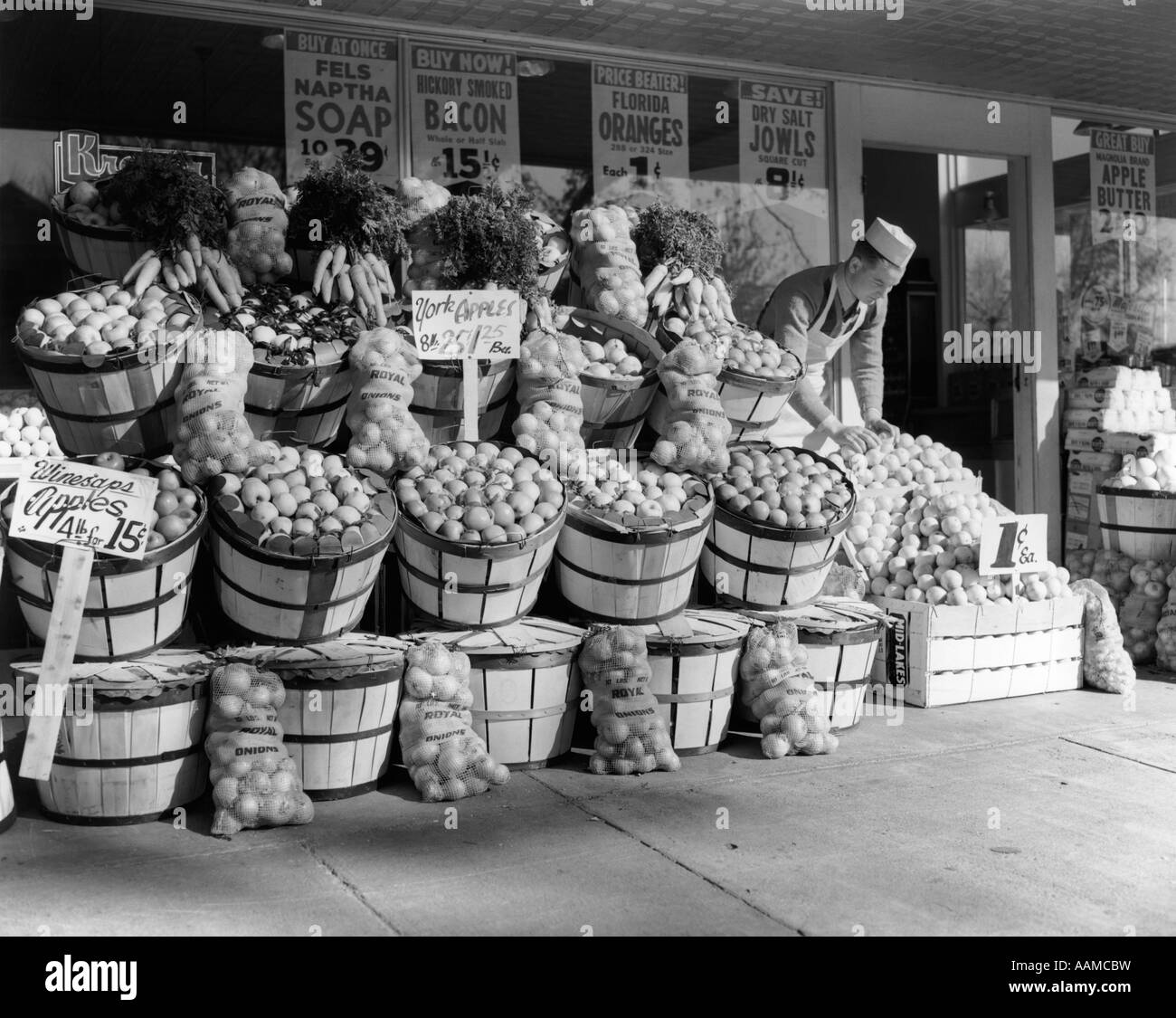 1940 GROCER EN BOUCHON BLANC TABLIER & EXPOSANT DES FRUITS SUR TROTTOIR EN FACE DE L'ÉPICERIE Banque D'Images