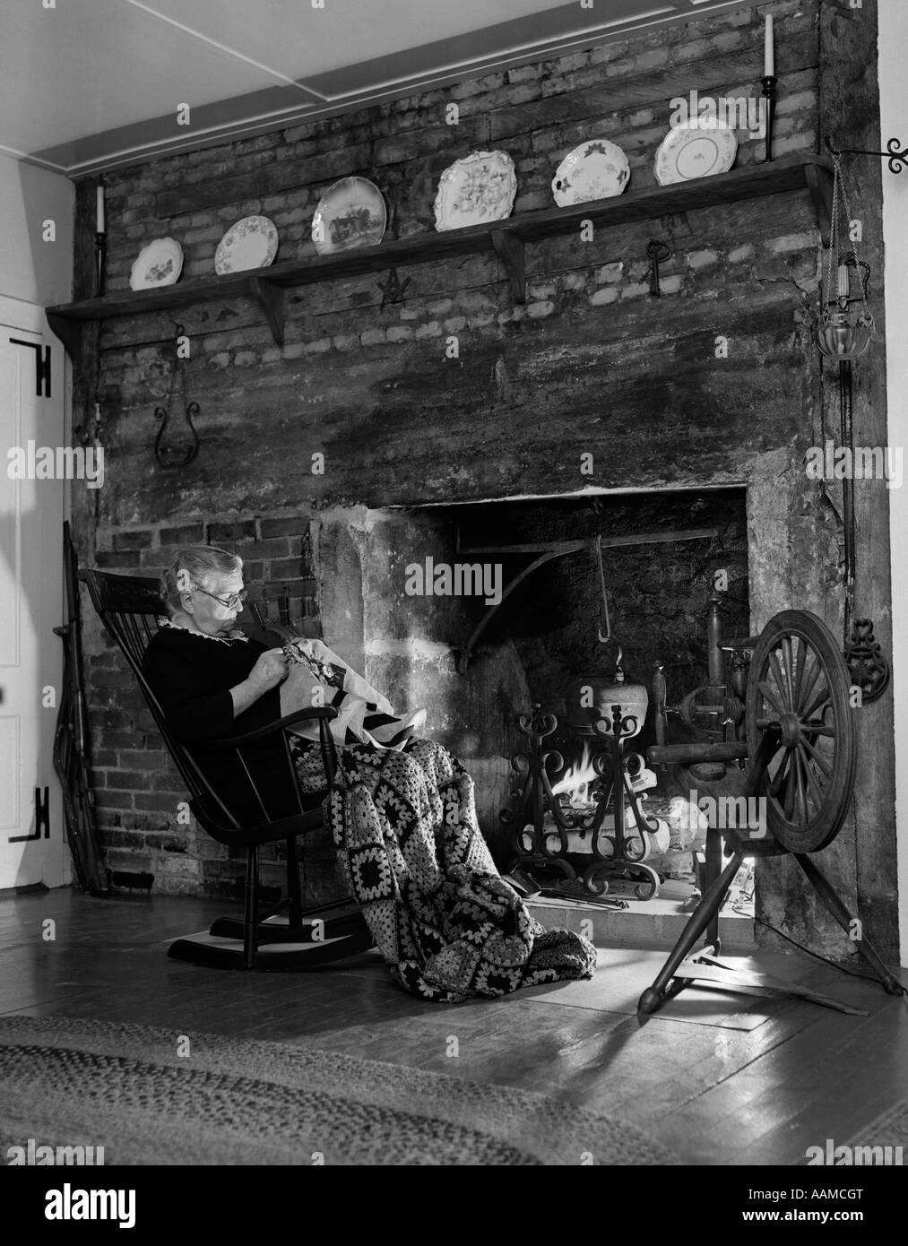 1950 femme âgée EN COUVRE-CULASSE EN FACE DE CHEMINÉE FAISANT LA COUTURE Banque D'Images
