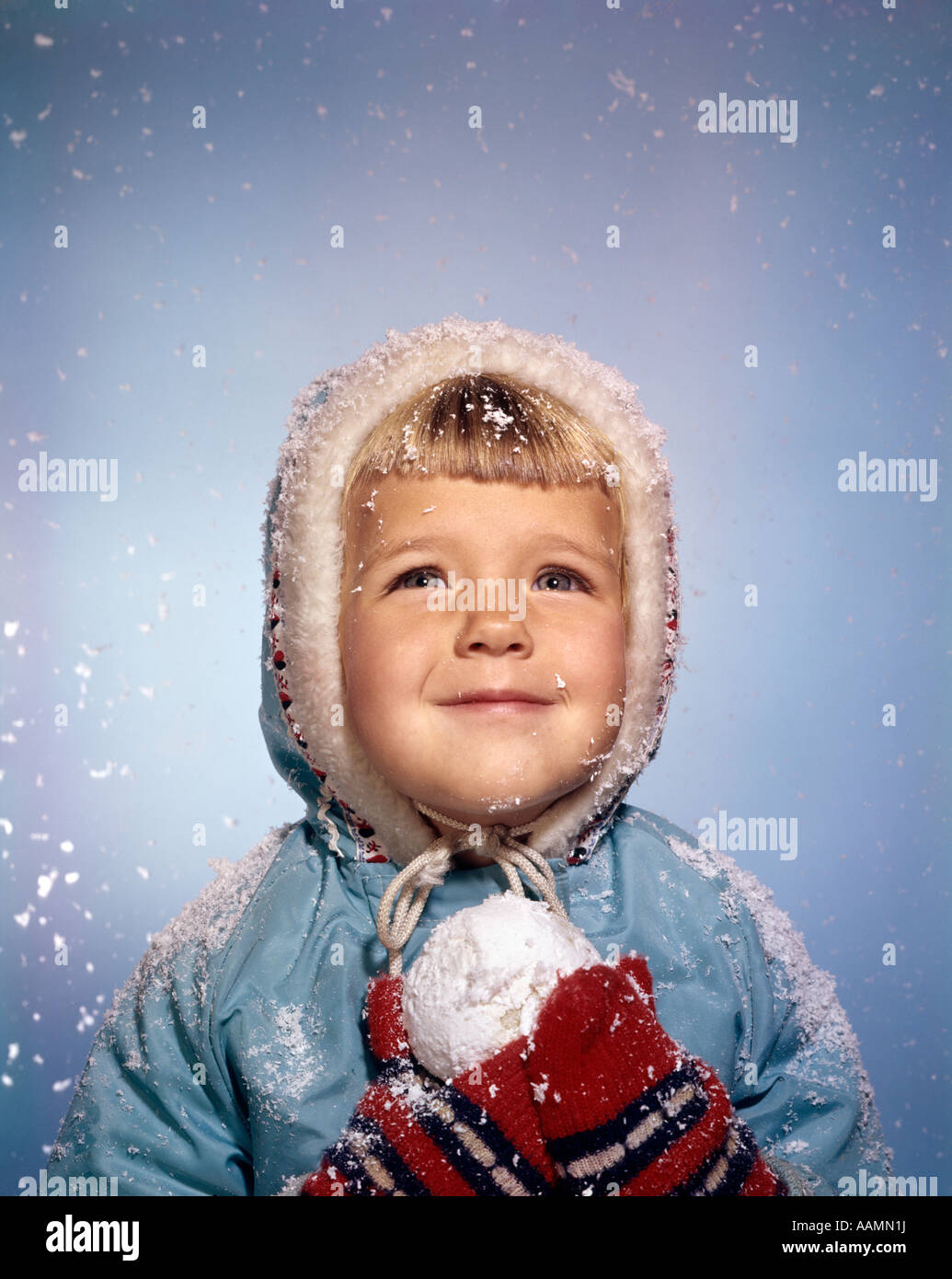 1960 LITTLE GIRL HOLDING SNOW BALL LOOKING UP AT SKY ROUGE SOURIANT BLANC MITAINES KNIT HAT VESTE BLEUE RETRO Banque D'Images