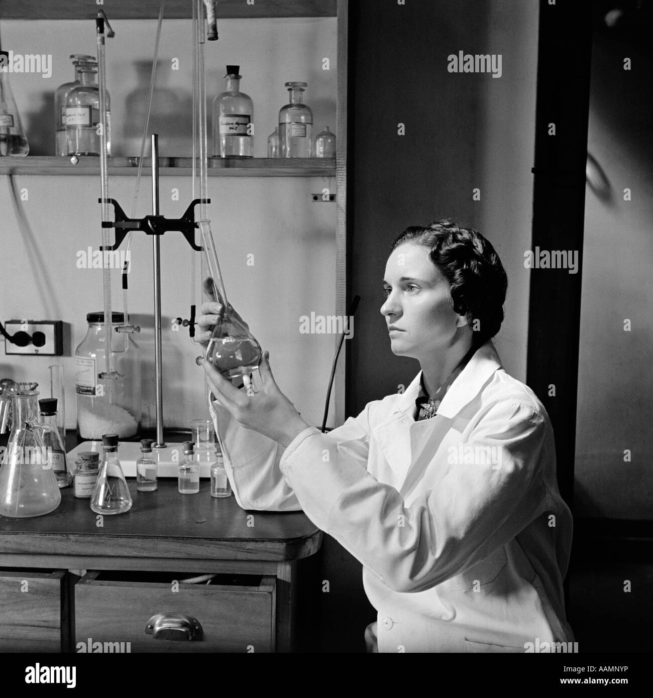 Années 1930 Années 1940 FEMME SCIENTIFIQUE IN LAB COAT HOLDING UP ET DE L'EXAMEN DU LIQUIDE BÉCHER Banque D'Images