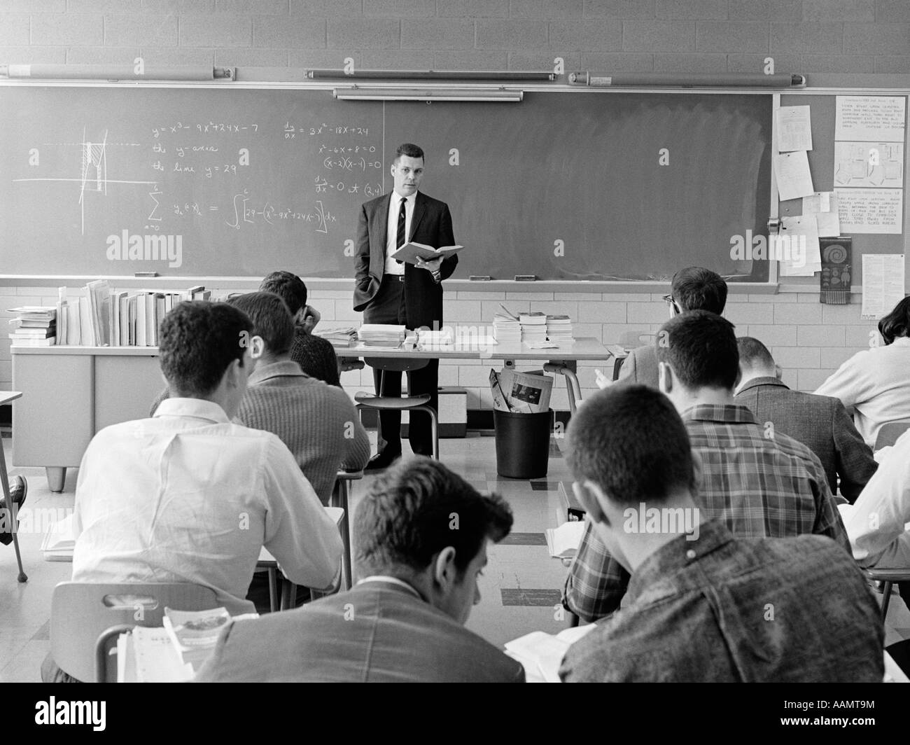 1960 VUE ARRIÈRE DE CLASSE AVEC L'ENSEIGNANT debout derrière un bureau avec LIVRE À LA MAIN EXPLIQUANT L'ÉQUATION SOUS FORME GRAPHIQUE SUR TABLEAU NOIR Banque D'Images
