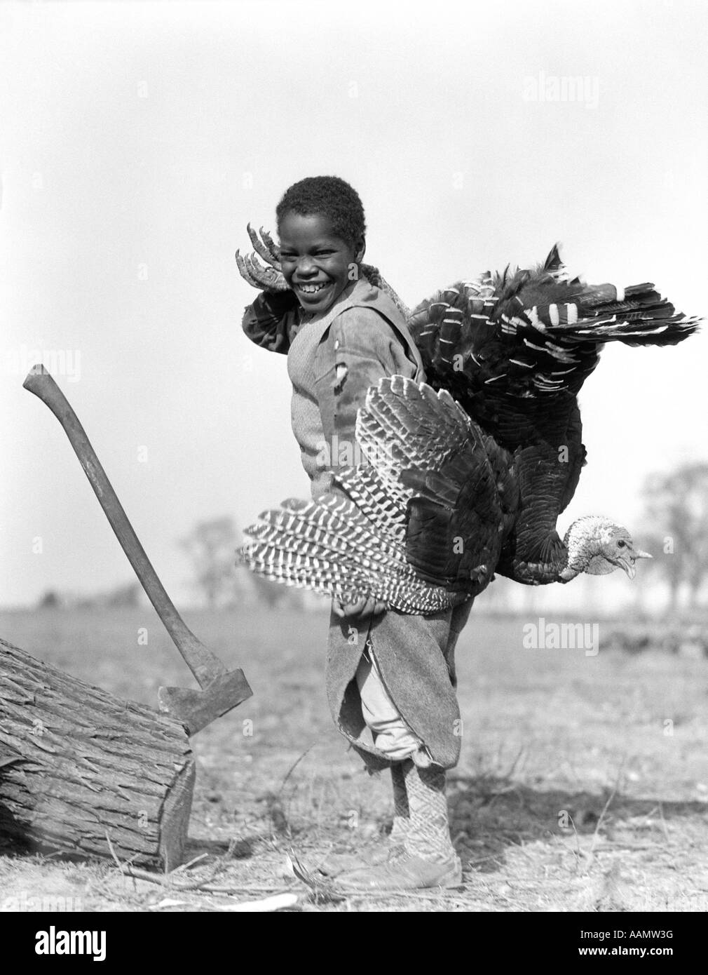 Années 1920 SMILING AFRICAN AMERICAN BOY EN KNICKERS LOOKING AT CAMERA TENAIT UNE TURQUIE COMITÉ PERMANENT PAR UN JOURNAL AVEC UNE HACHE Banque D'Images