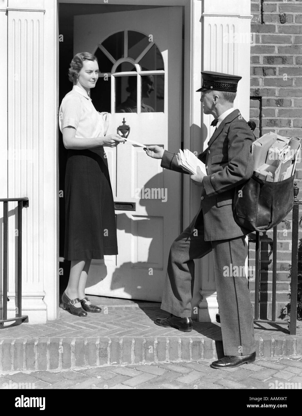 1930 POSTMAN DONNANT UNE LETTRE À UNE FEMME DANS L'embrasure D'UNE MAISON EN BRIQUE COLONIALE Banque D'Images