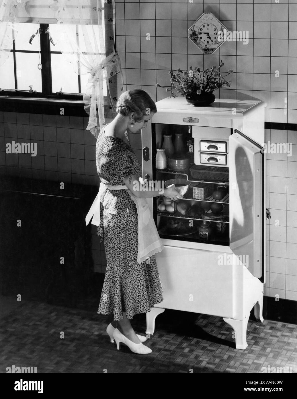 Années 1920 Années 1930 Femme blonde en robe de soirée en soie TABLIER HIGH HEELS PRENDRE PLAT DE CUISINE RÉFRIGÉRATEUR ÉLECTRIQUE Banque D'Images