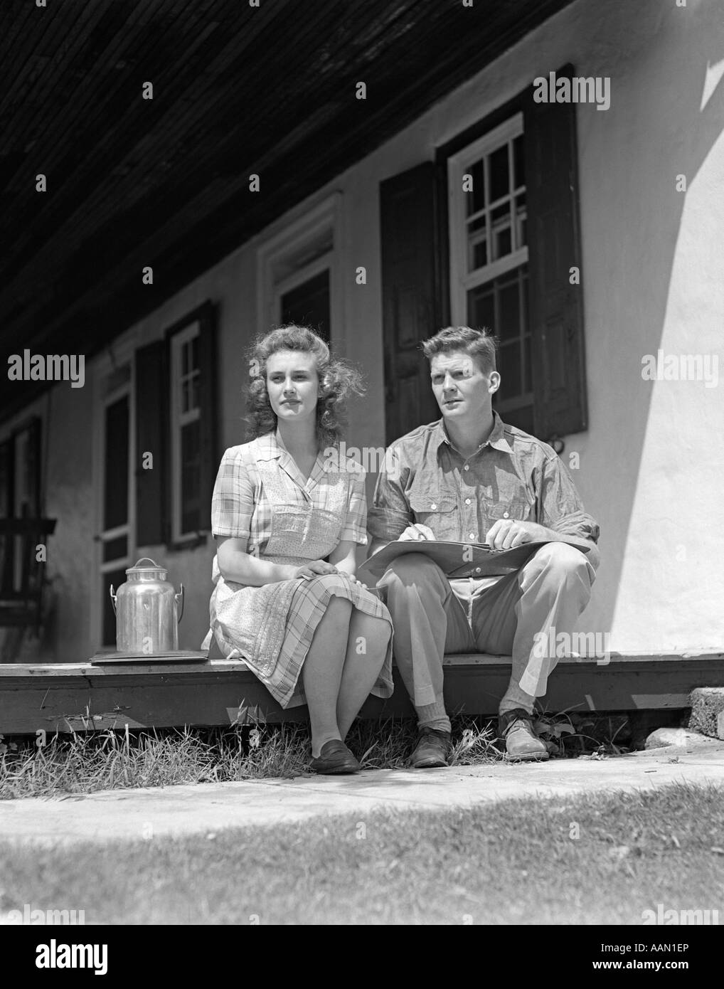 1940 HOMME FEMME COUPLE ASSIS À LA MAISON FERME PORCHE DE CÔTÉ GRAND LIVRE LES GENOUX de l'homme petite robe à carreaux Femme du réservoir de lait Banque D'Images