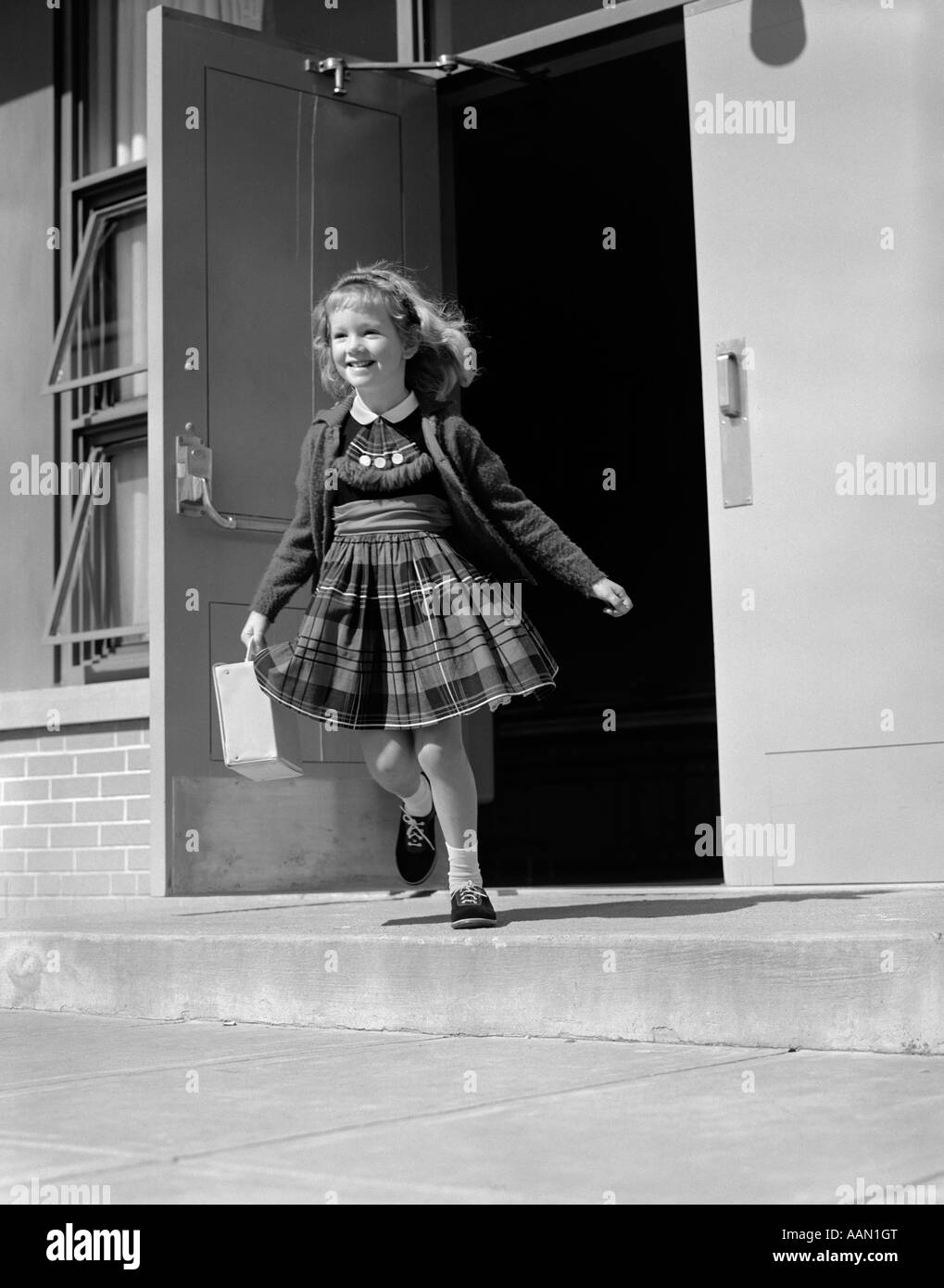 Années 1950 Années 1960 fille blonde COURT PORTE AVANT DU BÂTIMENT DE L'ÉCOLE TENUE LUNCH BOX OU SAC pull robe à carreaux SMILING Banque D'Images