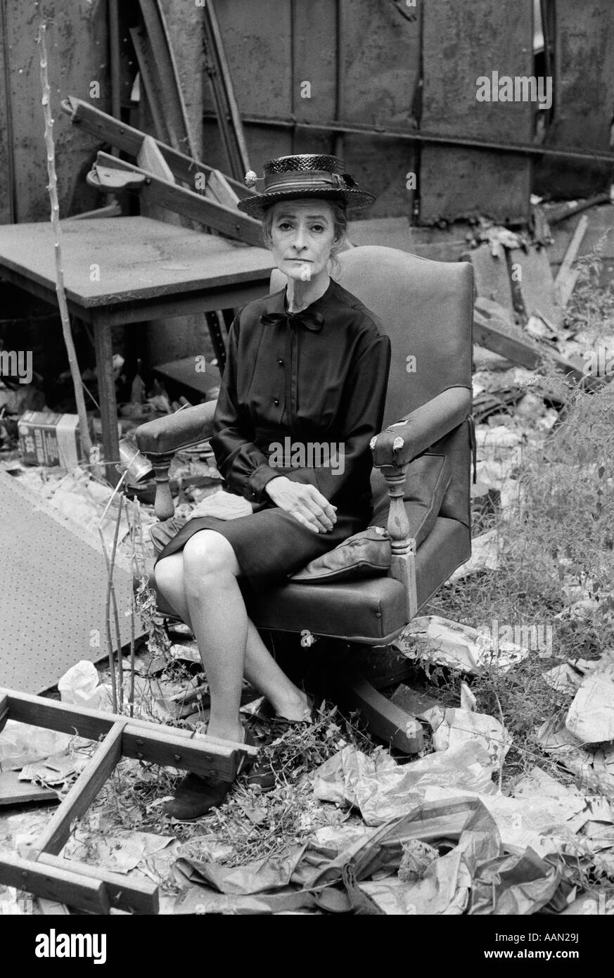 Des années 70, vieille femme en robe et chapeau assis sur fauteuil pivotant en cuir parmi les ruines & CORBEILLE AVEC EXPRESSION MOROSE Banque D'Images