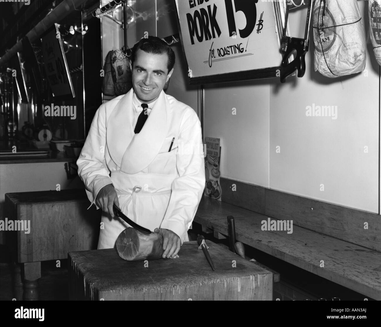 Années 1930 Années 1940 SMILING MAN SLICING BOUCHER ROULEAU DE JAMBON DANS UNE BOUCHERIE LOOKING AT CAMERA Banque D'Images