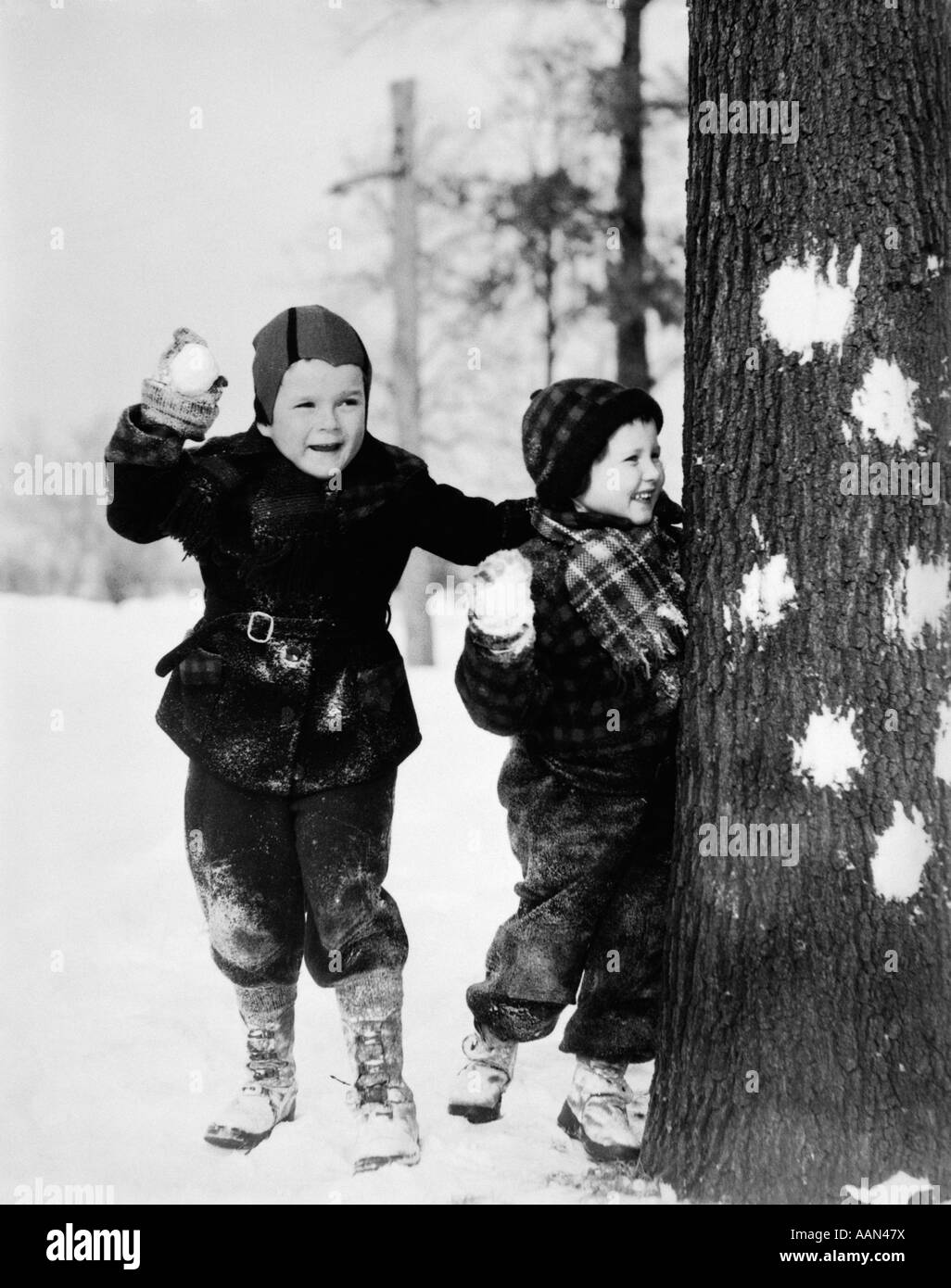 Années 1920 Années 1930 SMILING BOY AND GIRL JOUER DANS LA NEIGE CACHE DERRIÈRE TREE AVEC DES BOULES DANS LA MAIN Banque D'Images