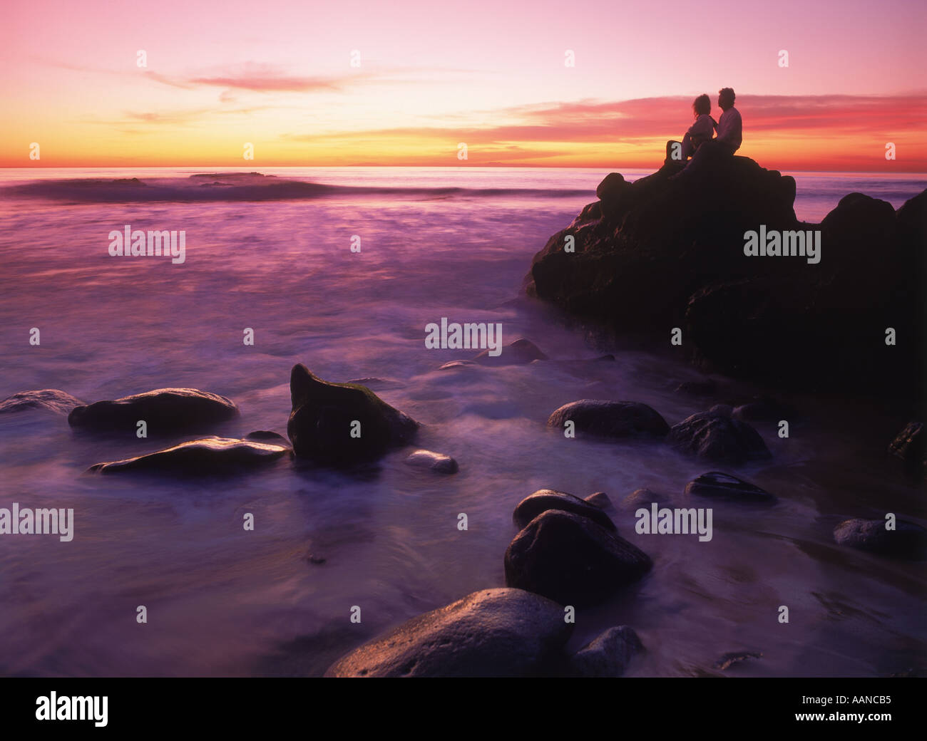 Couple assis sur côte rocheuse au-dessus de vagues au coucher du soleil en Californie Banque D'Images