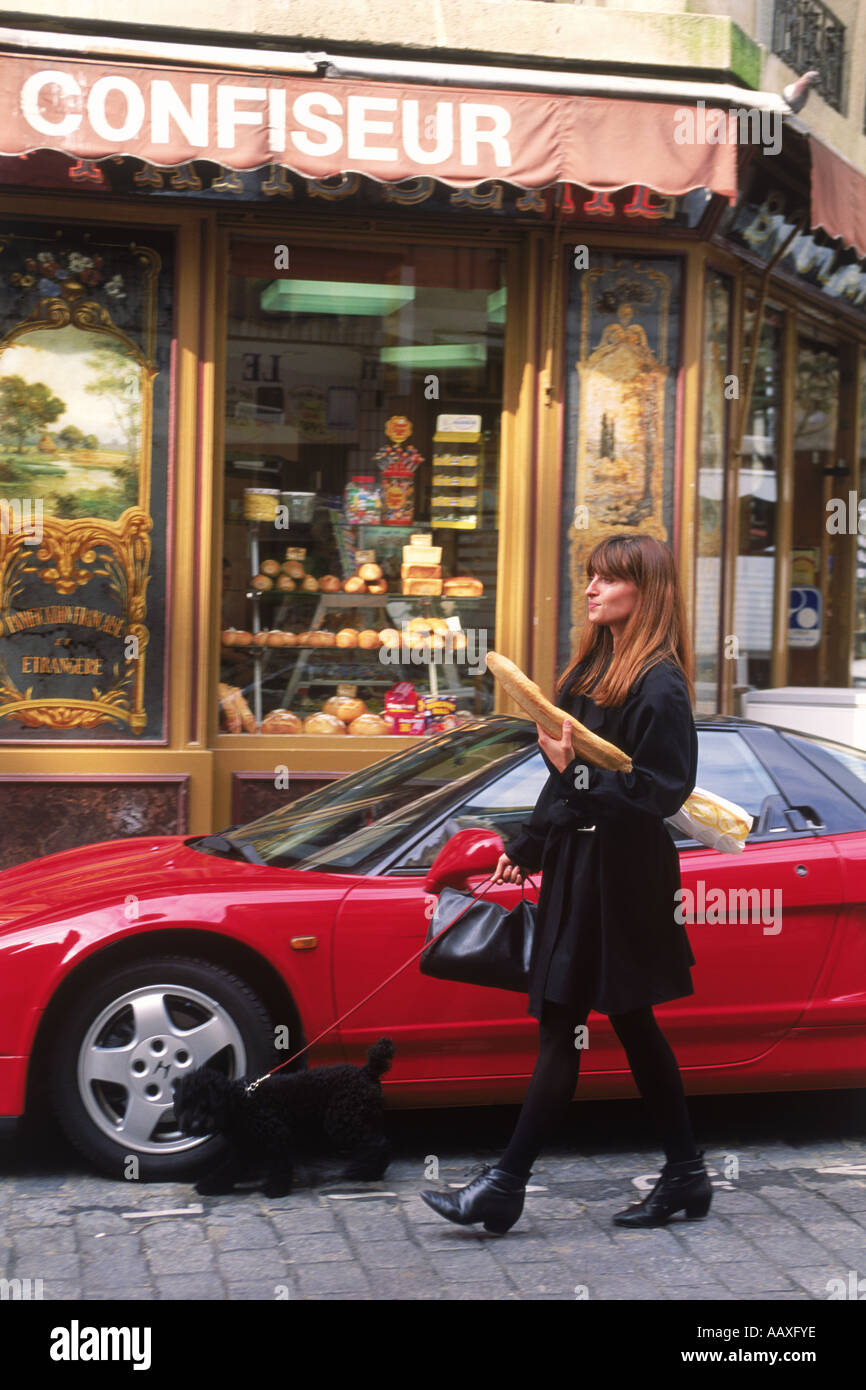 Femme à Paris laissant boulangerie baguette avec passant voiture sport rouge avec caniche français sur cobblestone street Banque D'Images