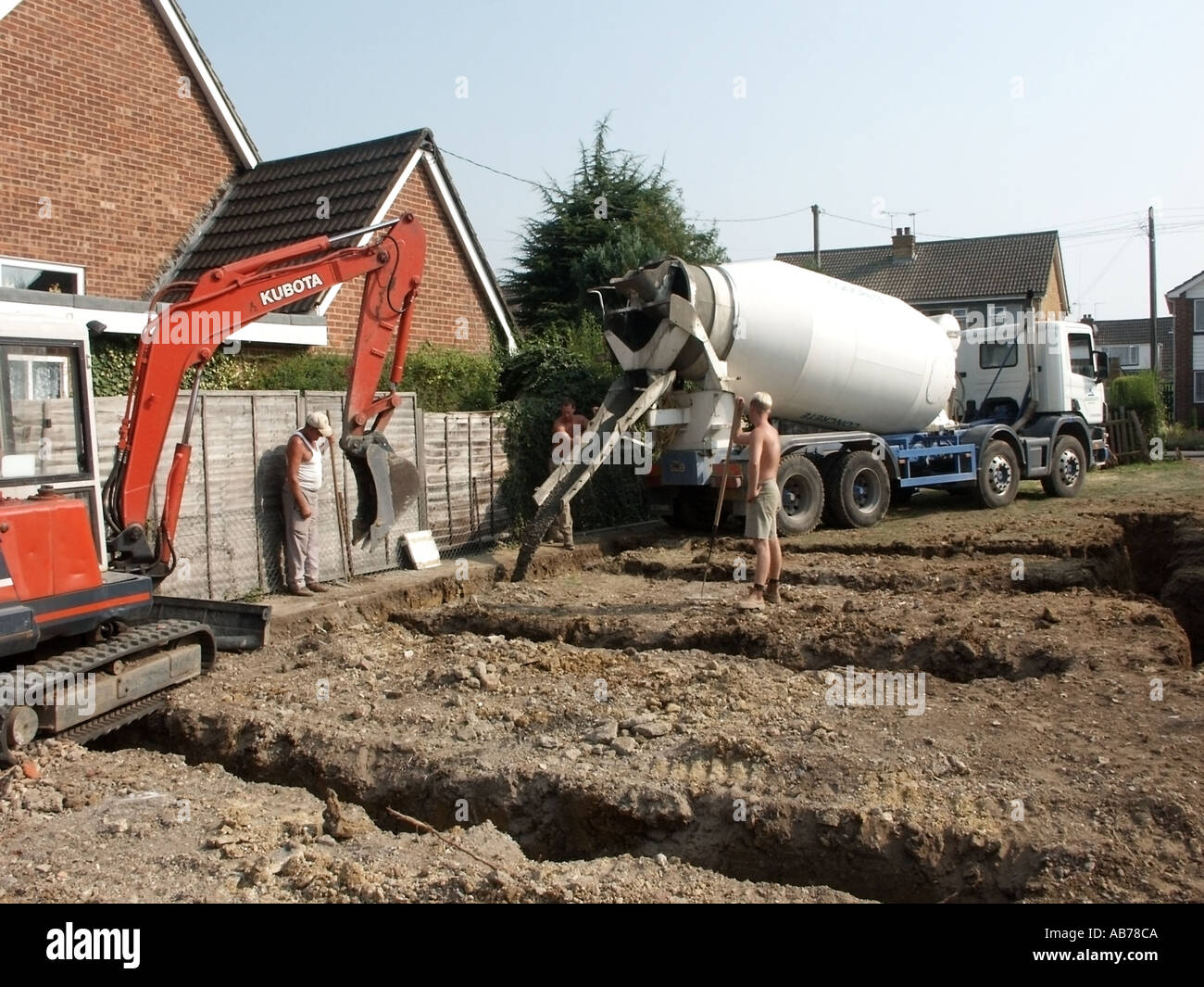 Les travaux en cours des camions de livraison de béton en remplissage de tranchées de fondation nouvelle maison individuelle avec mini pelle sur chantier plot Essex UK Banque D'Images
