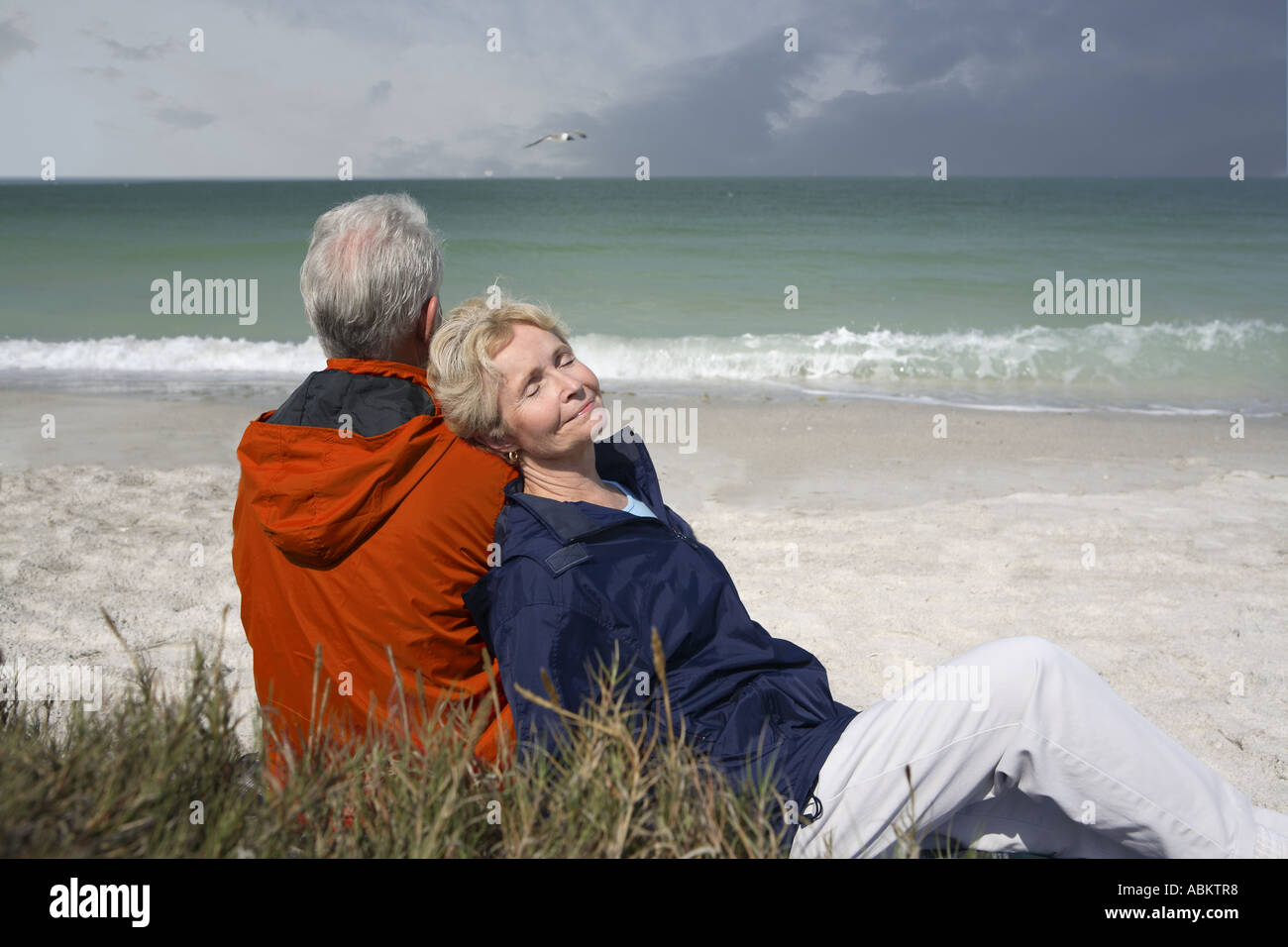 Homme d'âge moyen et la femme assise sur la plage portant l'orange et le bleu marine coupe-vent Banque D'Images