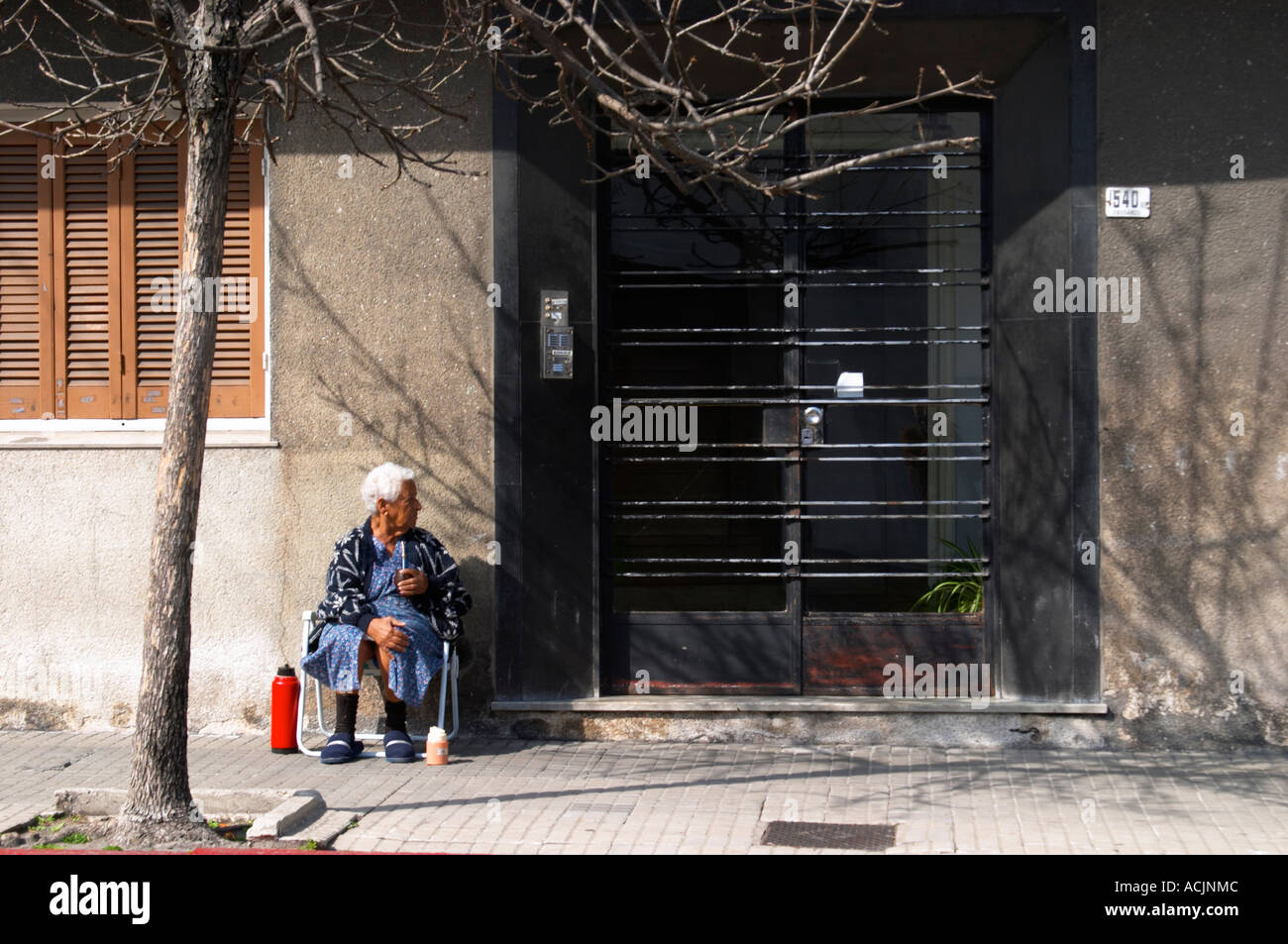 Une vieille femme assise sur le trottoir de boire une tisane mate, avec un ballon d'eau chaude thermos rouge en face d'une porte de fer noir porte. Montevideo, Uruguay, Amérique du Sud Banque D'Images