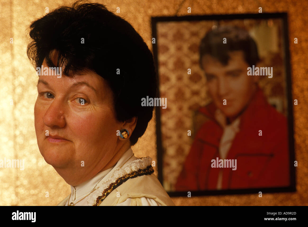 Elvis Presley fan, Mme Valérie Constable portant des boucles d'oreilles de portrait Elvis, portrait d'Elvis sur le mur du salon derrière elle.1990s Londres Royaume-Uni Banque D'Images