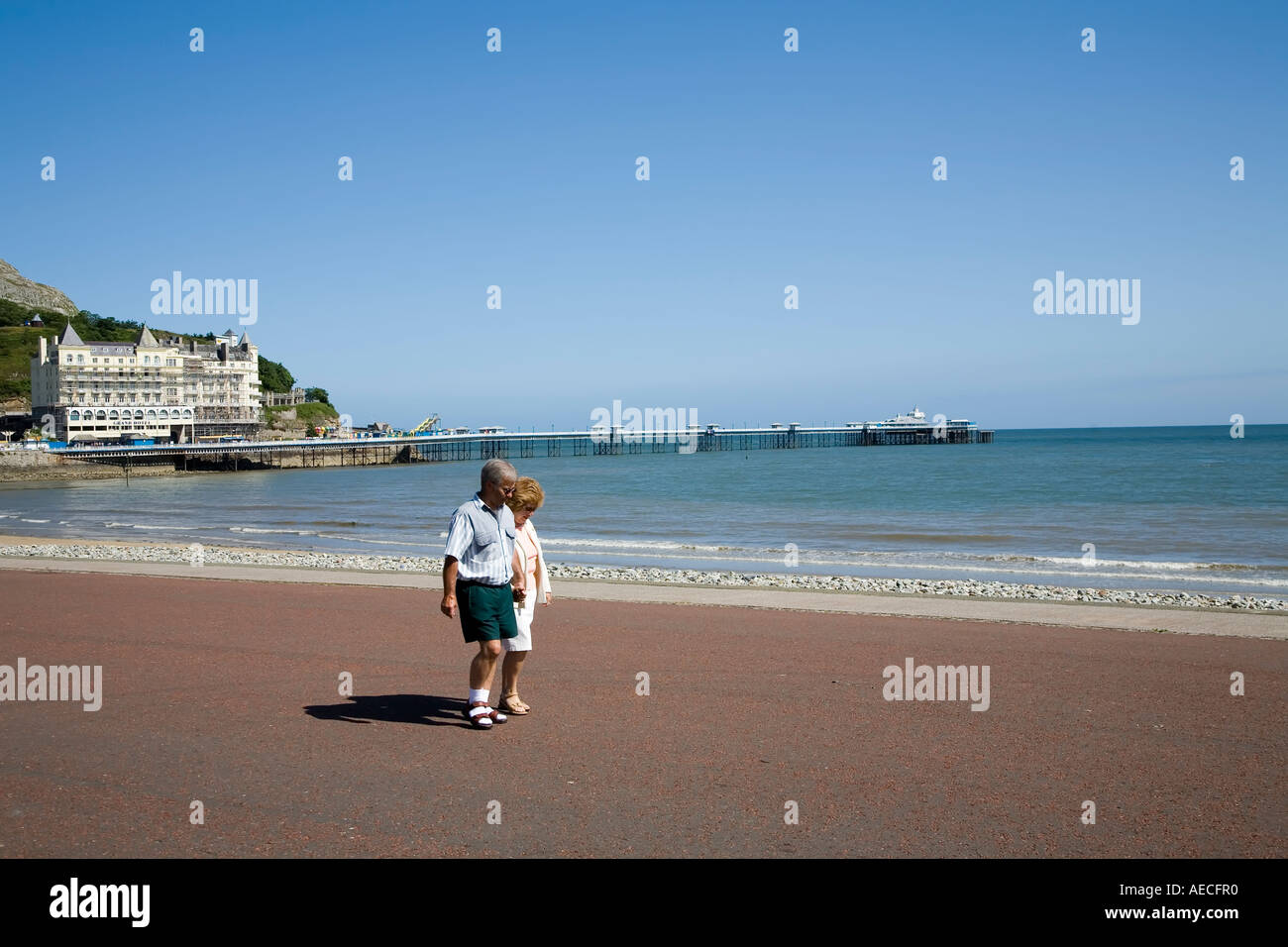Couple d'âge moyen marche sur la promenade avec le grand hôtel et jetée victorienne dans la distance Llandudno Galles UK Banque D'Images