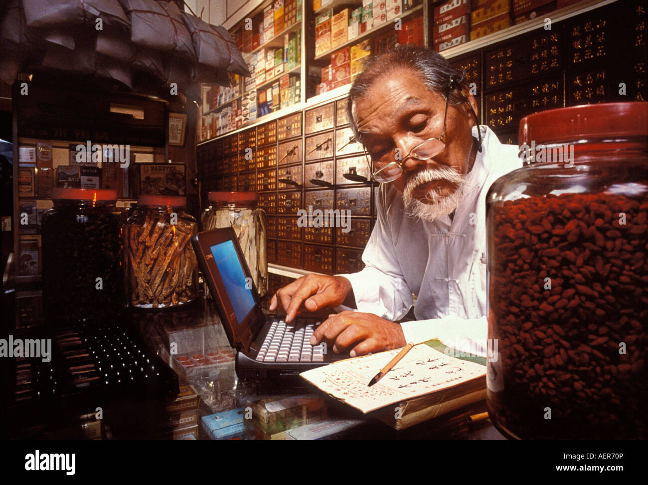 Un pharmacien chinois traditionnel dans une mise d'apothicaire Singapour ses formules anciennes d'un ordinateur portable Banque D'Images