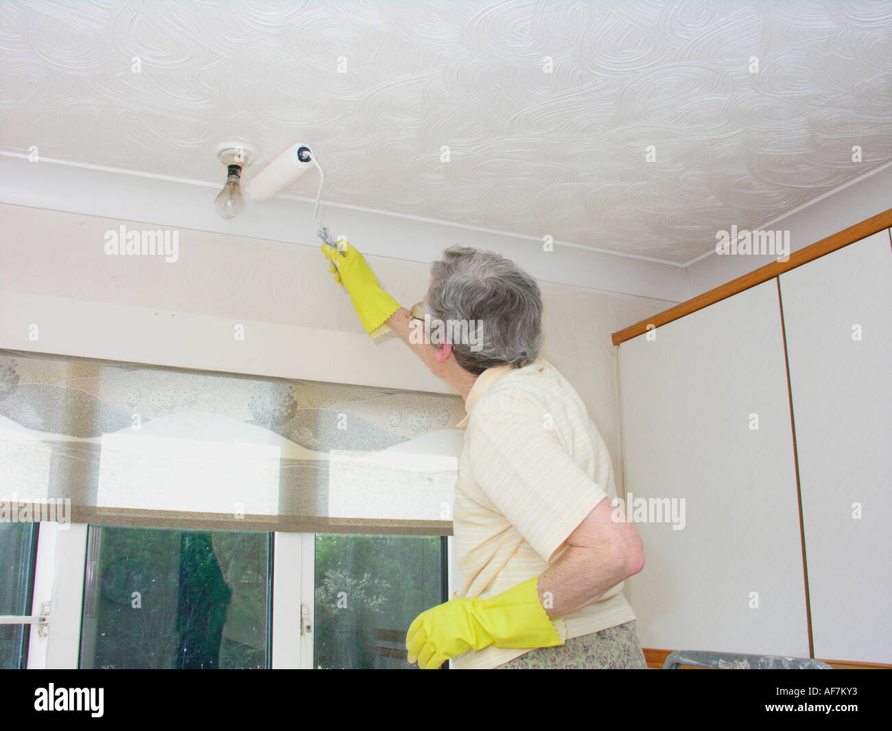 Femme âgée de la décoration et de la peinture du plafond avec un rouleau à peindre et d'avoir à se pencher sur les raccords fixes dans la cuisine Banque D'Images