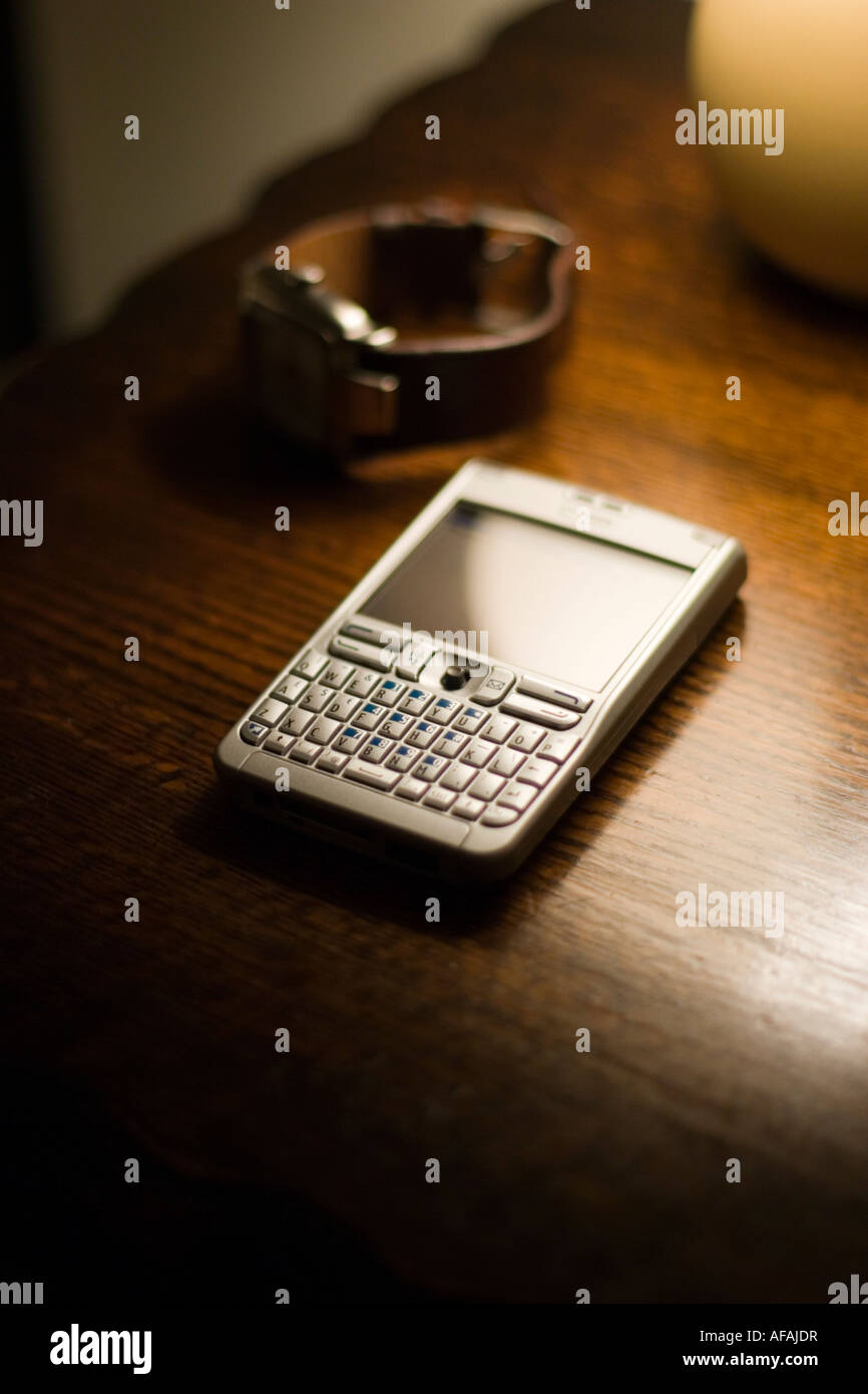 Un Nokia E61 smartphone sur une table antique poli avec un mens watch avec un bracelet en cuir brun éclairé par une petite lampe Banque D'Images