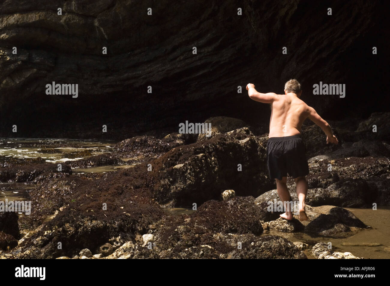 Homme d'âge moyen vu de dos portant un maillot de rock à l'autre dans un bassin de marée entre de gros rochers Banque D'Images