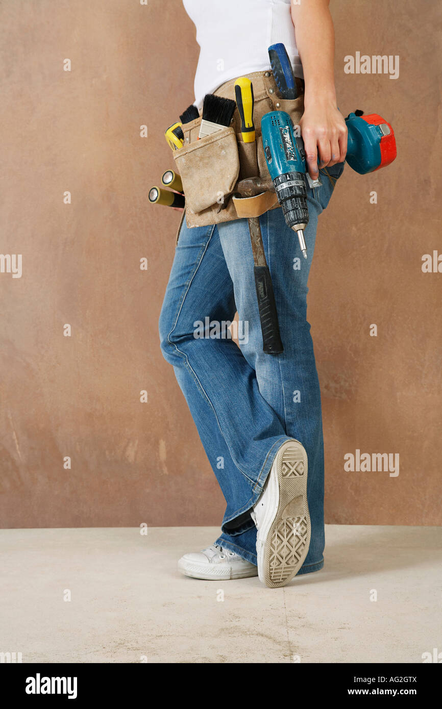 Femme avec toolbelt et percer leaning against wall, low section Banque D'Images